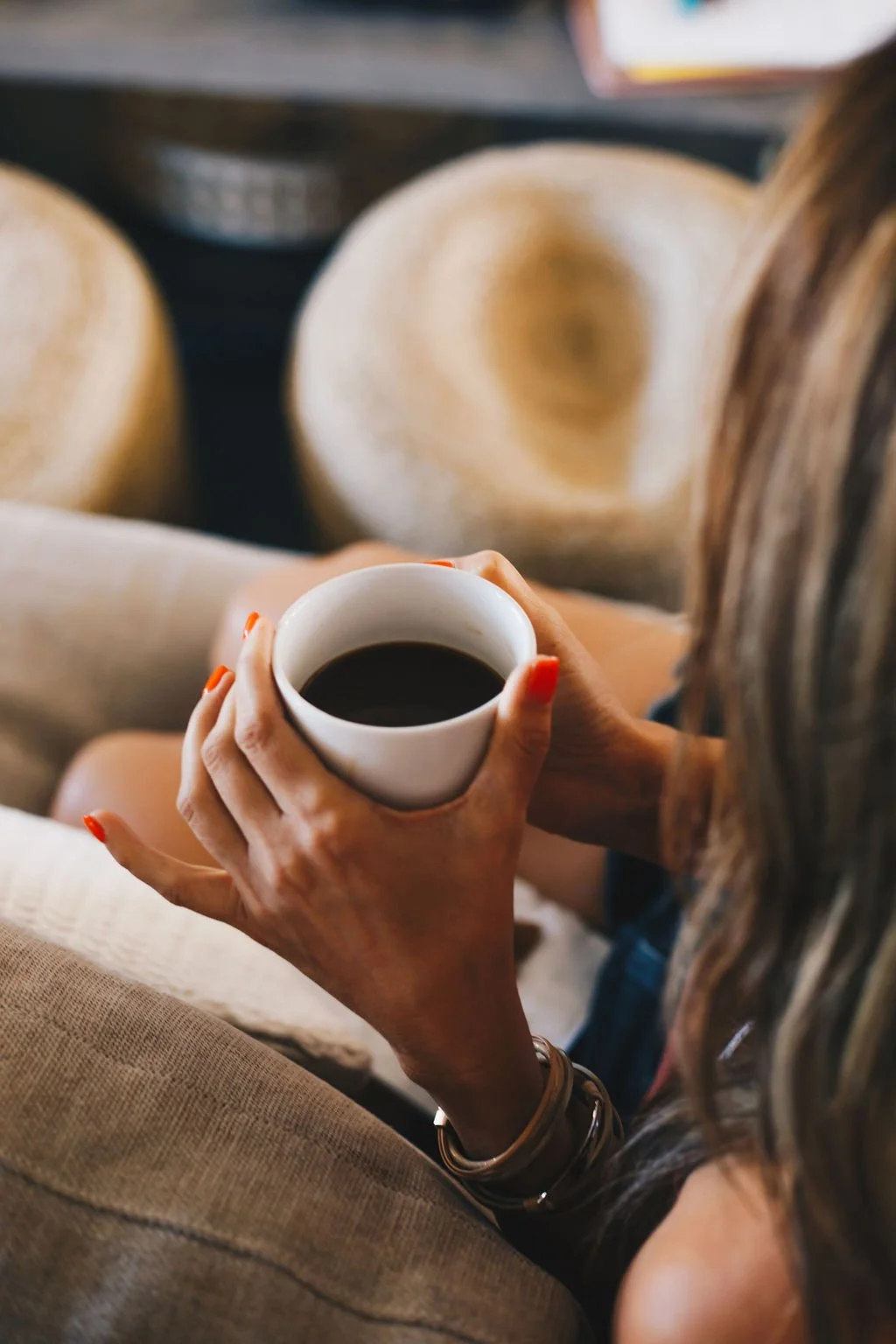 Person holding a white mug of black coffee with red nail polish and a bracelet, seated on a beige couch with woven hats in the background.