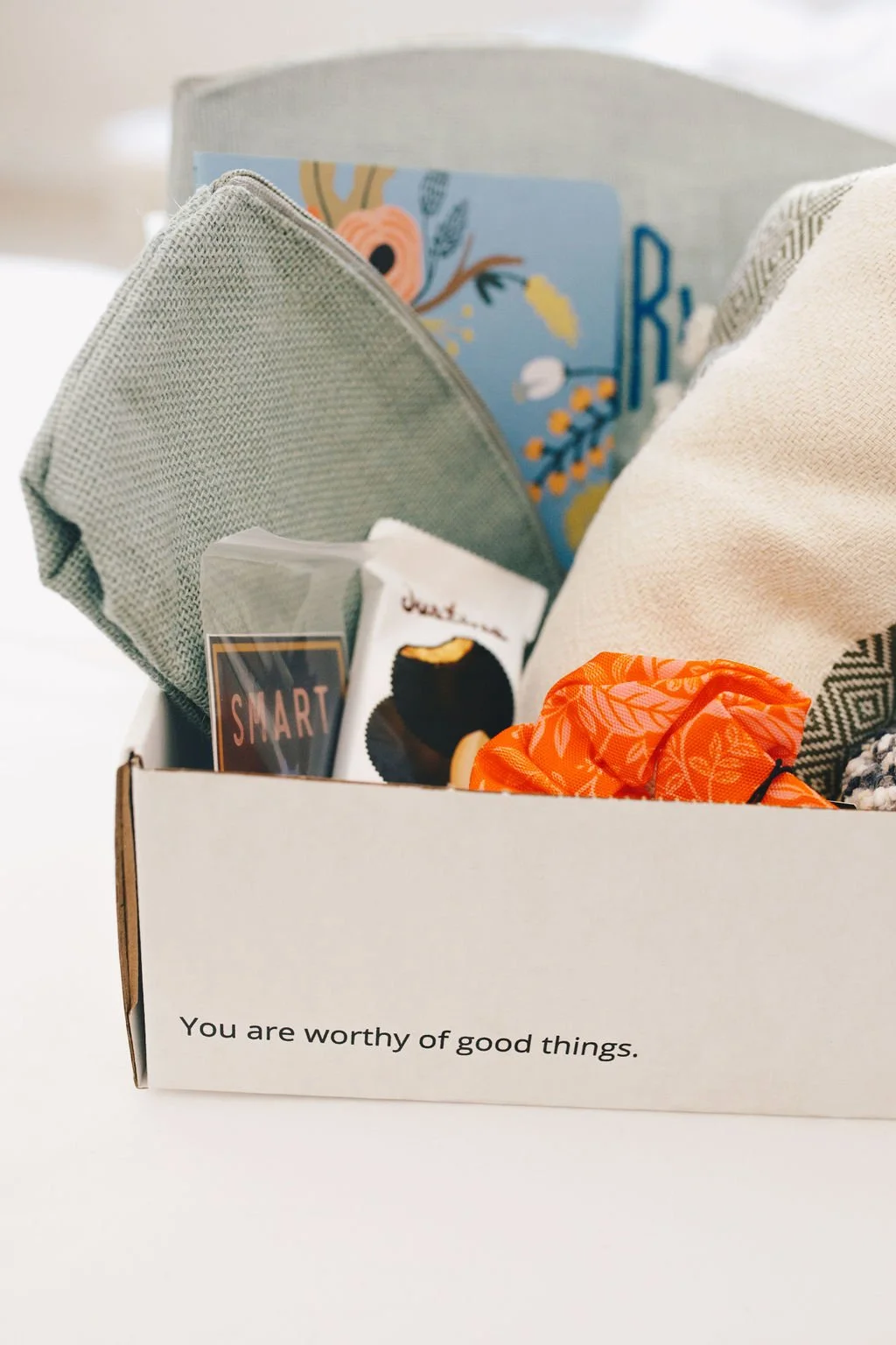 A box with the message "You are worthy of good things" contains various items including a gray cloth, a face mask, a magazine, a snack, and an orange cloth or cosmetic pouch.