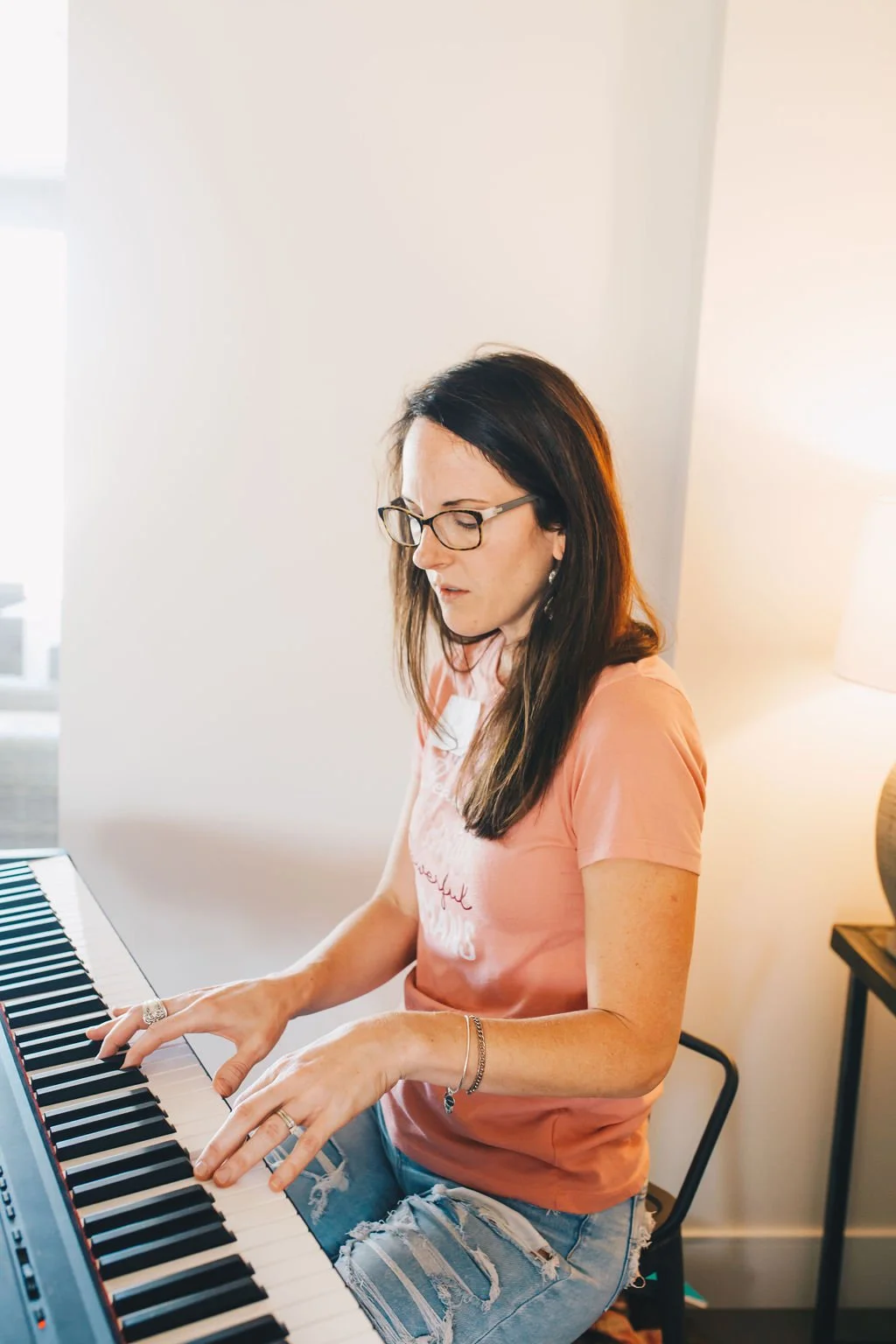 A woman with long dark hair, glasses, and earrings playing a digital piano in a bright room, wearing a pink T-shirt and ripped jeans.