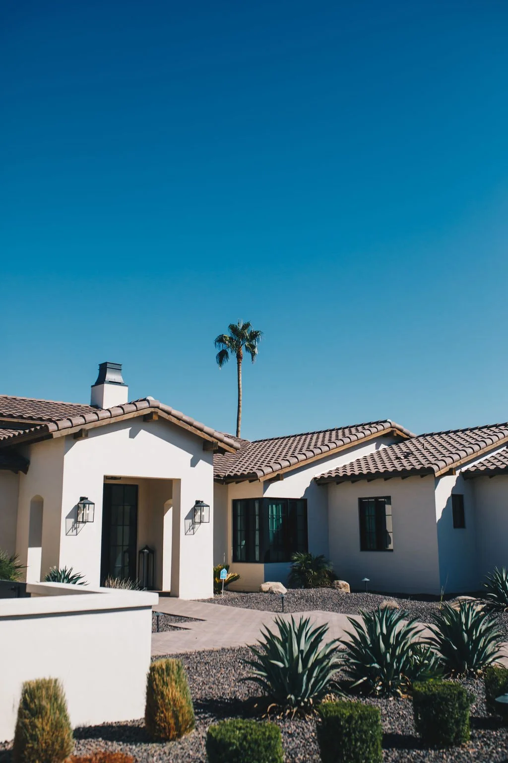 A house with a beige stucco exterior, brown tiled roof, and black window frames, surrounded by desert plants, with a clear blue sky and a tall palm tree in the background.