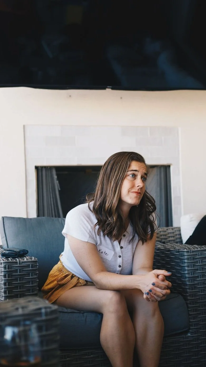 A woman with shoulder-length brown hair and a white shirt sitting with her hands clasped, looking to her right, in a living room with a fireplace and wicker furniture.