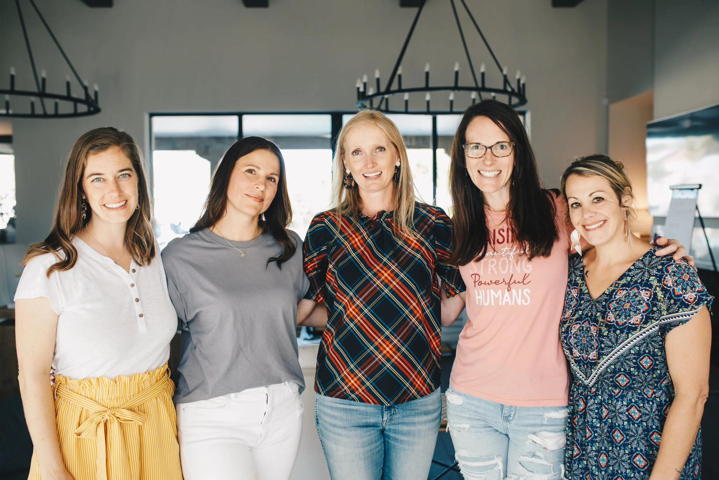 Group of five smiling women standing together indoors, with three large chandeliers hanging above and windows in the background.