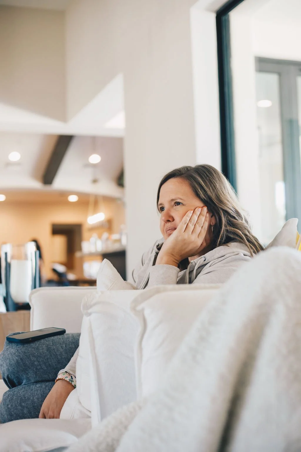 A woman sitting on a couch appears bored or pensive, with her hand resting on her face, in a bright, modern interior with large windows.