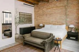 Bedroom with a bed, white bedding, and two nightstands with lamps, a bench at the foot of the bed, a full-length mirror, and a window with a brick wall backdrop.