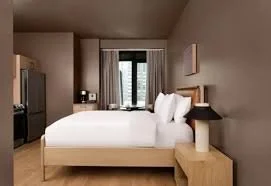 A neatly made bed in a hotel room with white linens, a nightstand with a lamp, and a window with curtains showing city buildings outside.