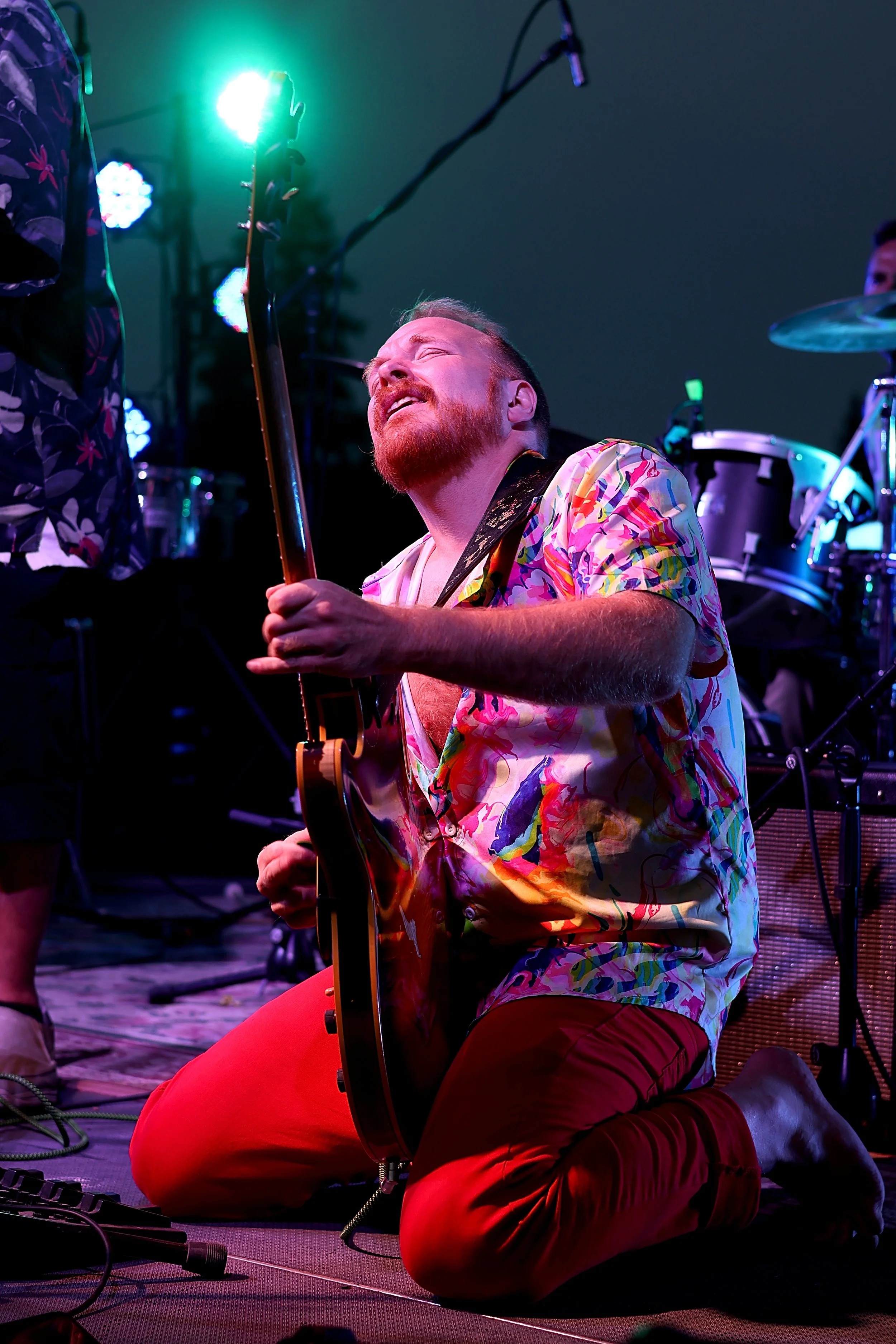 Musician kneeling on stage playing an electric guitar with a colorful shirt and red pants during a concert with colorful stage lights.