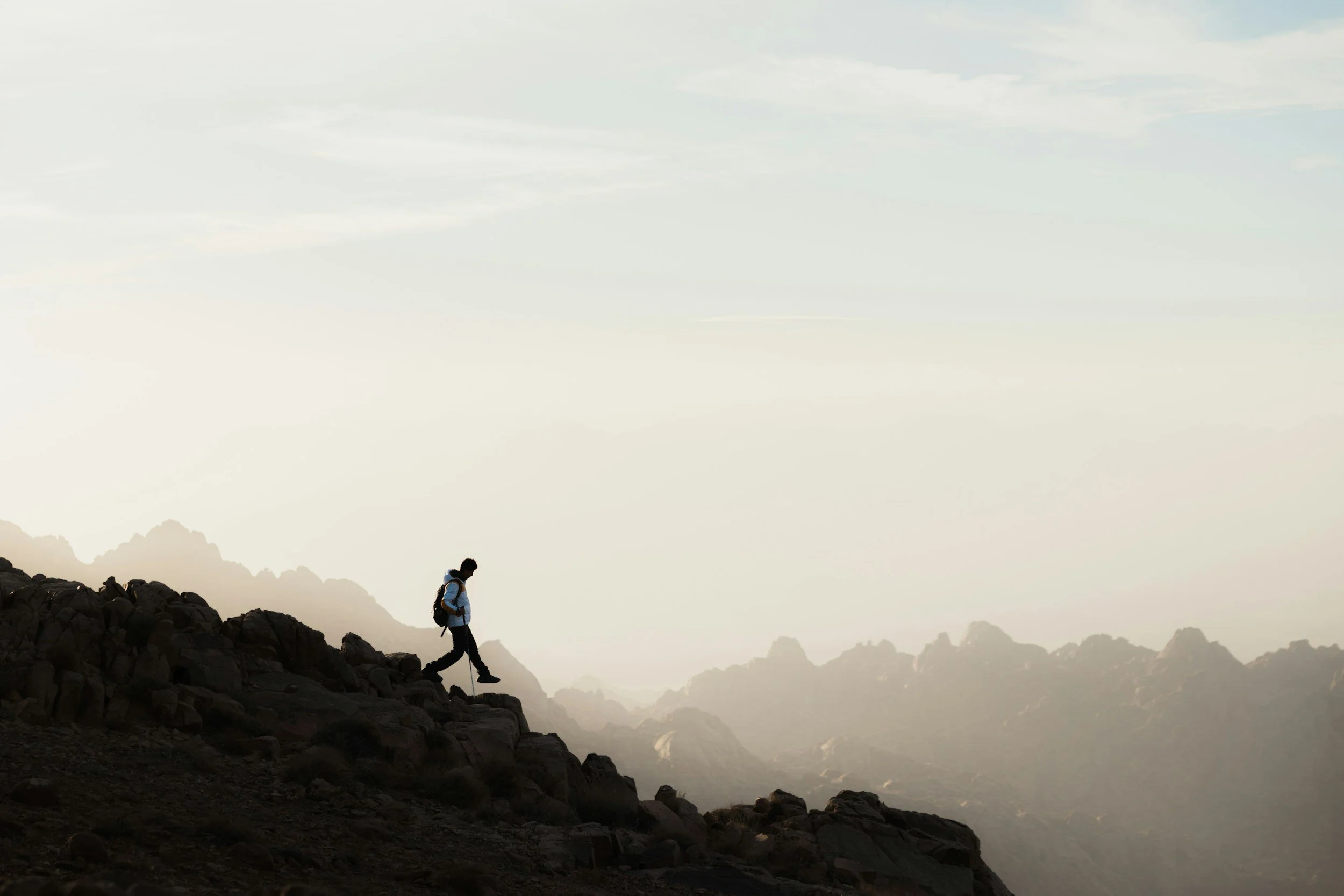 A hiker walking along a narrow mountain trail on a steep slope with a backpack, overlooking a valley with green fields and mountains under a partly cloudy sky.