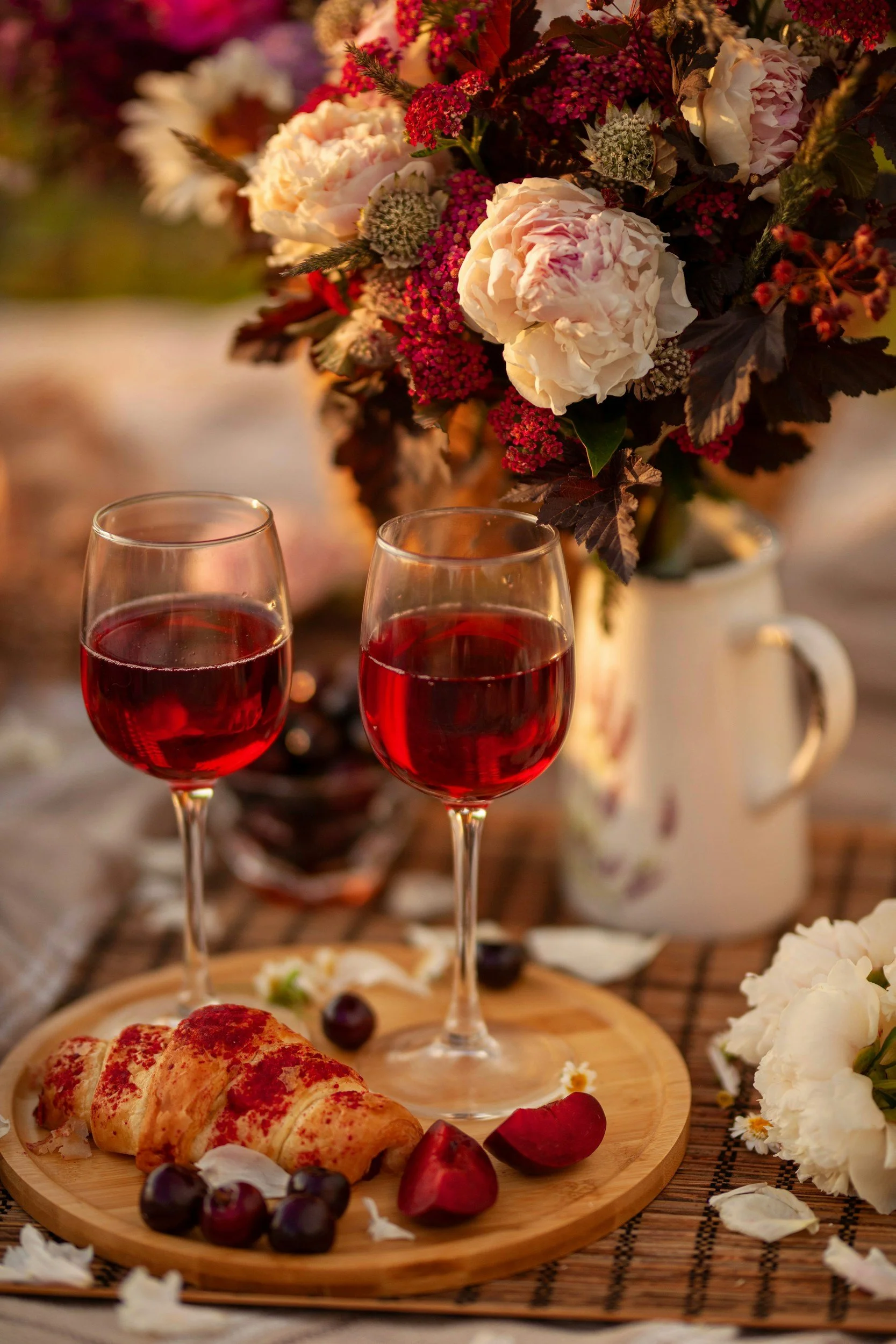A romantic table setting with two glasses of red wine, a croissant on a wooden plate, fresh cherries and rose petals, set against a backdrop of a large floral bouquet with pink and white flowers in a white pitcher.