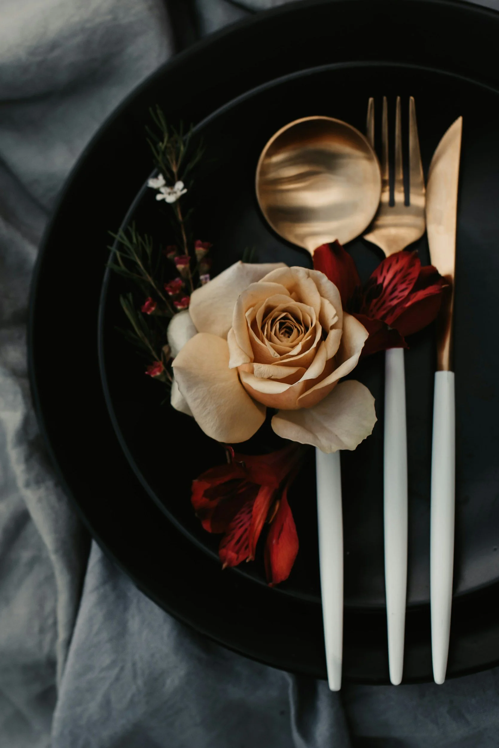 A black plate with a beige rose and red orchids, a gold spoon, fork, and knife are placed on the plate, with the utensils arranged around the flower. The plate is on a textured gray surface.