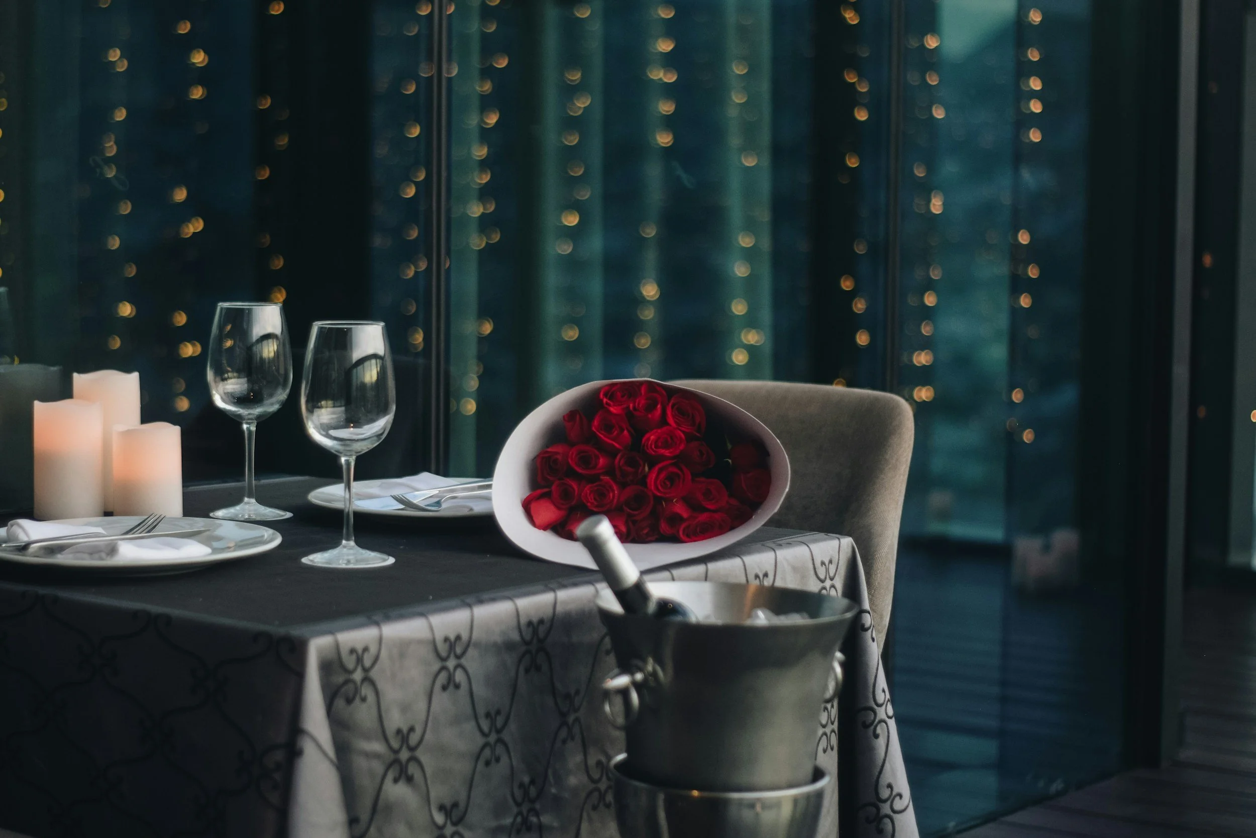 A romantic dinner setup with a table, candles, glasses, and a bouquet of red roses on a gray chair in front of a glass window with city lights outside.