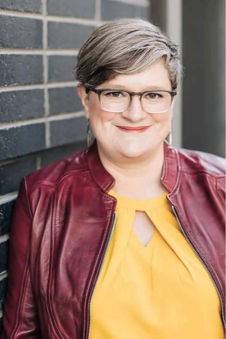 Close-up of a woman with short gray hair, wearing glasses, a yellow top, and a red leather jacket, smiling against a brick wall background.
