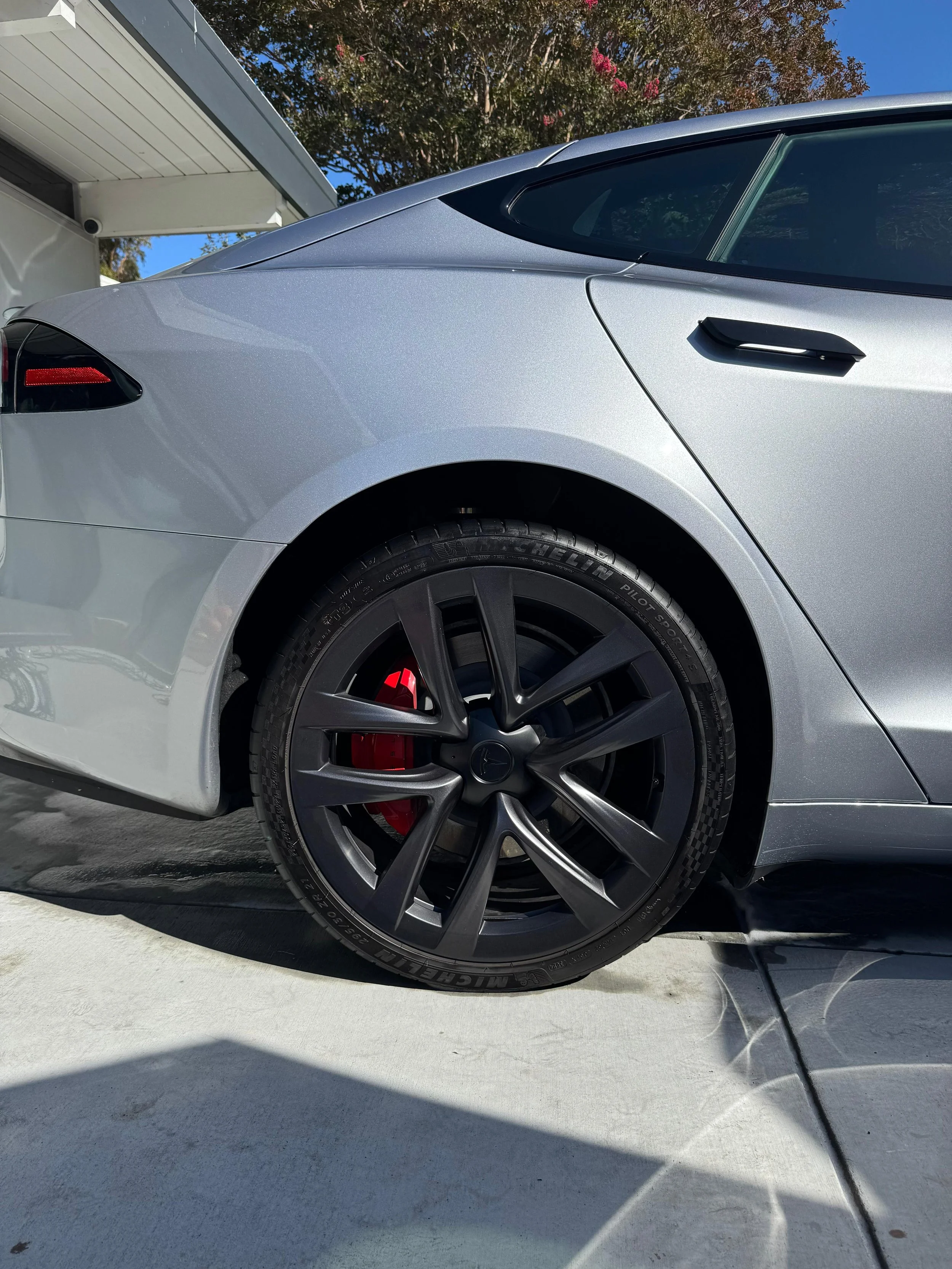 Close-up of the rear side of a silver Tesla Model S showing the rear wheel with black rim, red brake caliper, and a portion of the car's body and window, with a driveway and a house in the background.