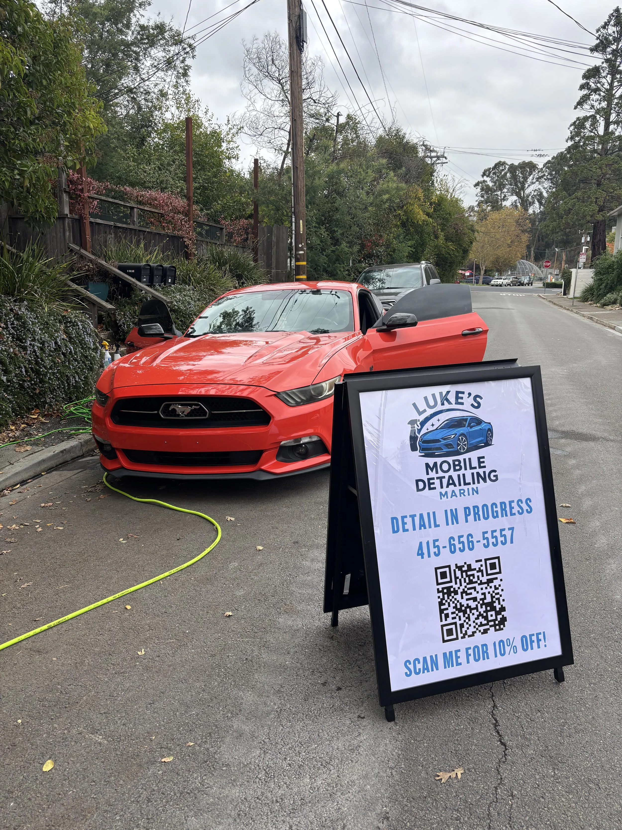 A red Ford Mustang is parked on the street during a car detailing service. A sign advertising Luke's Mobile Detailing in Marin with a phone number and QR code is positioned in front of the car.