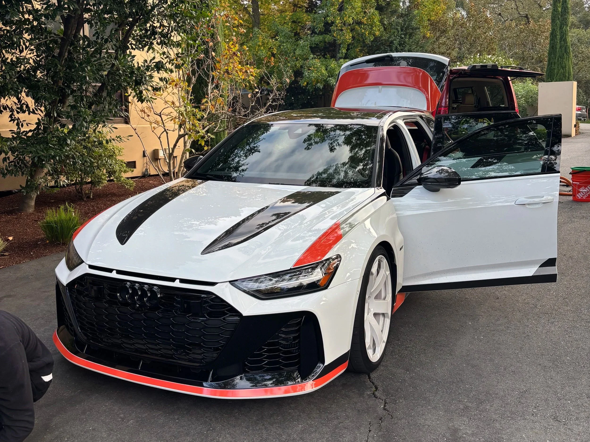 A white Audi sports car with black and red accents, positioned outside, with a red and black vehicle with an open trunk behind it, and trees and buildings in the background.