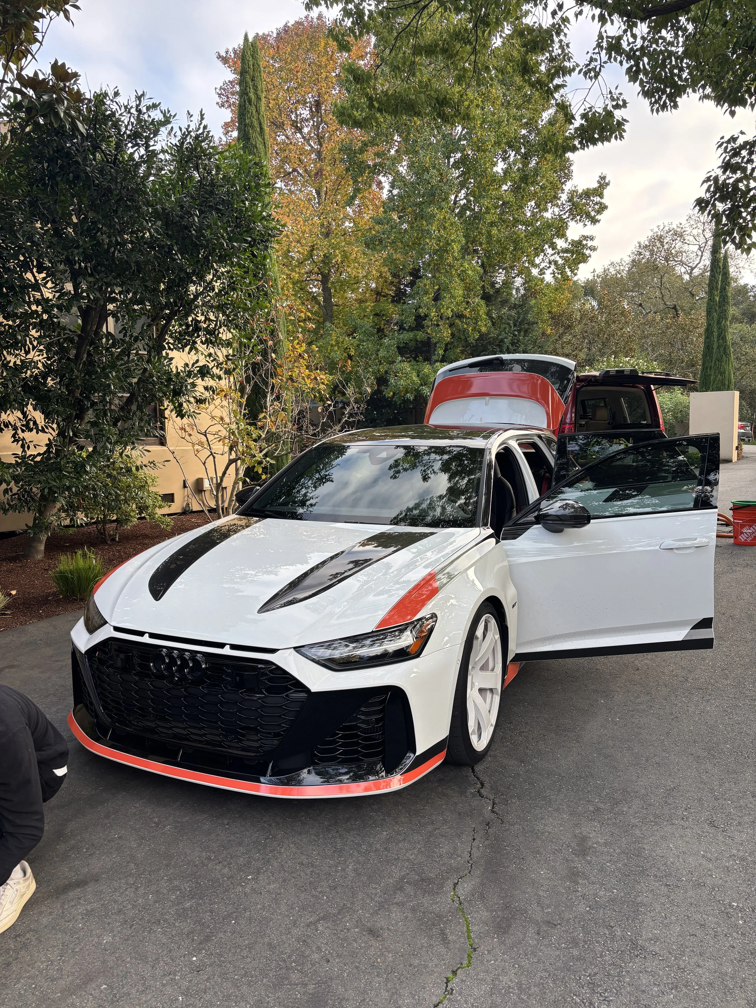A white sports car with black and red accents parked on a driveway next to trees and a building, with the front and rear doors open.