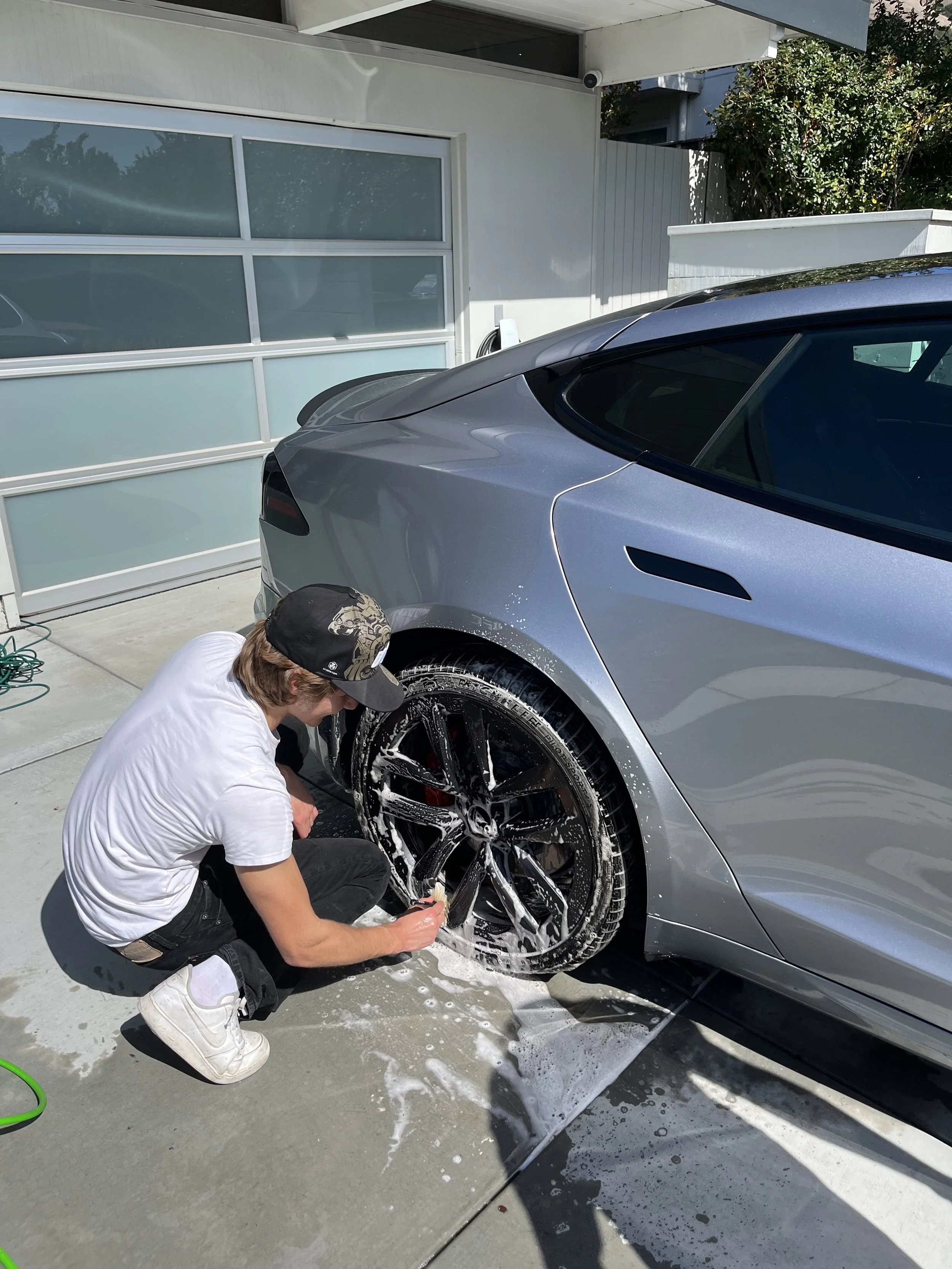 Person washing a grey sports car with soap and water on a driveway.