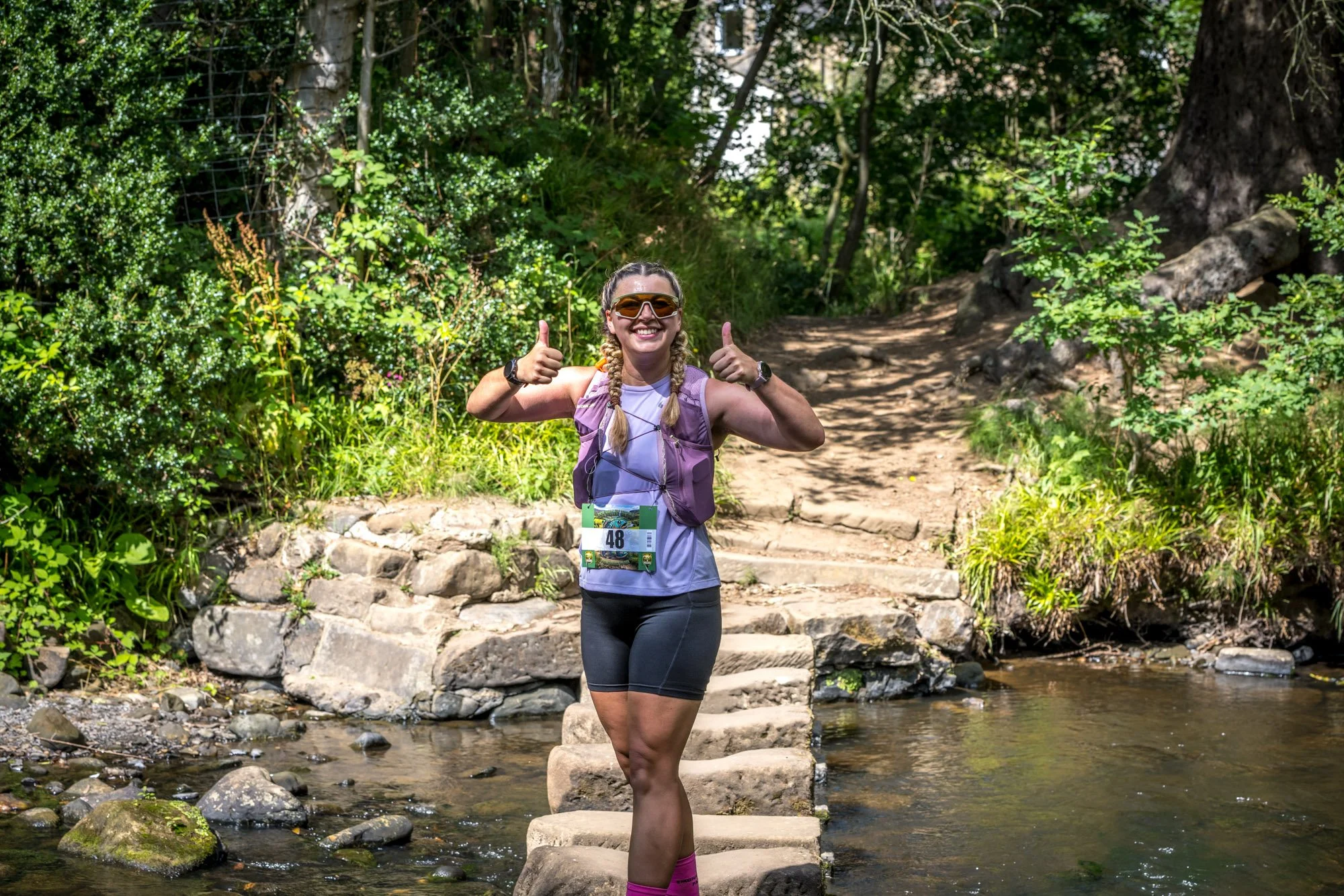 A woman wearing running gear and sunglasses is smiling and giving two thumbs up while crossing a small stream on stone steps in a wooded outdoor setting.
