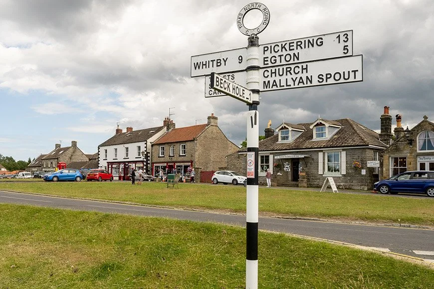 Street sign in a small town with buildings, cars, and people in the background, under cloudy skies.