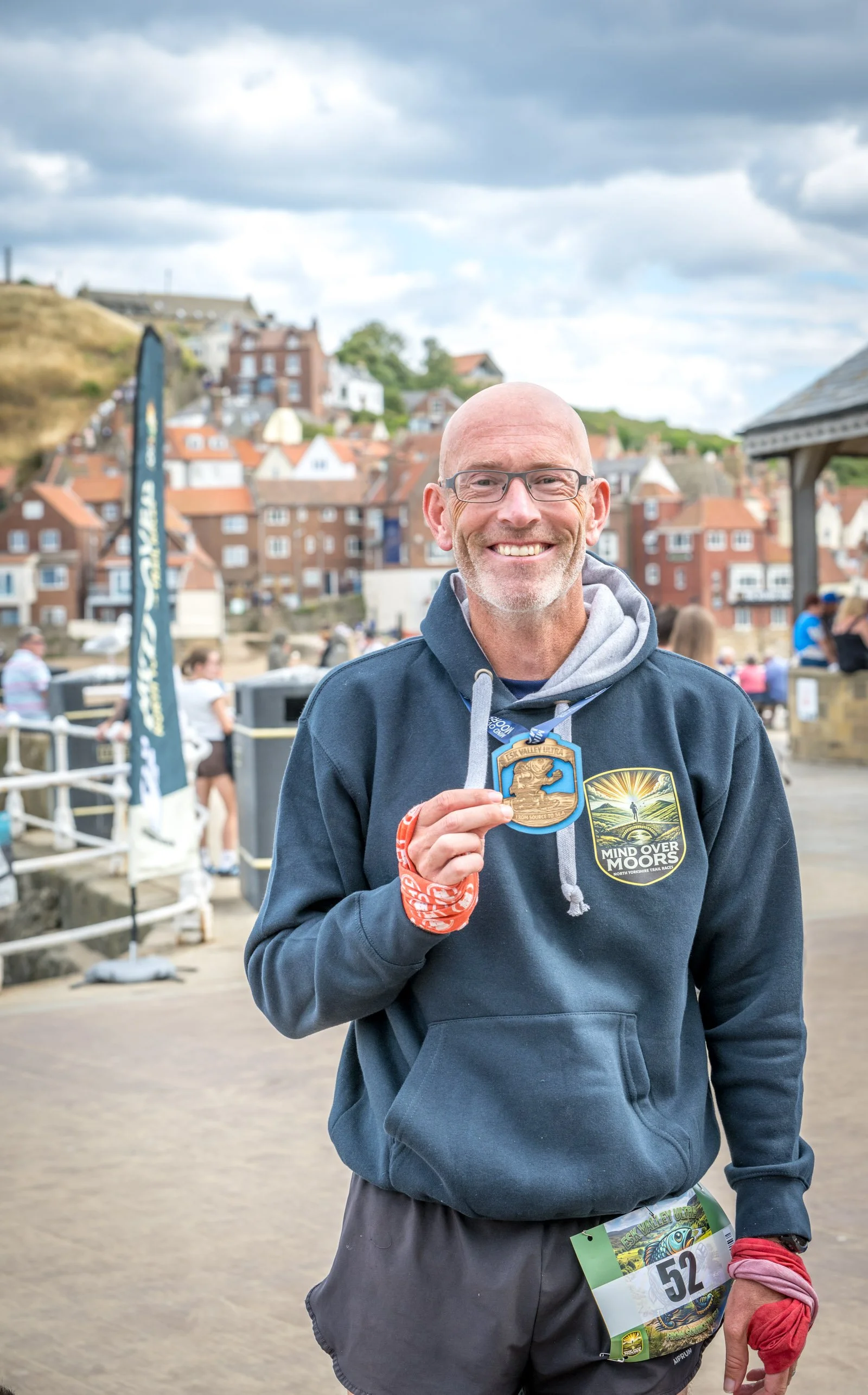 A smiling man with glasses, wearing a dark hoodie with a race badge and the number 52, holding a medal and a red band around his wrist, standing outdoors with people and colorful houses in the background.