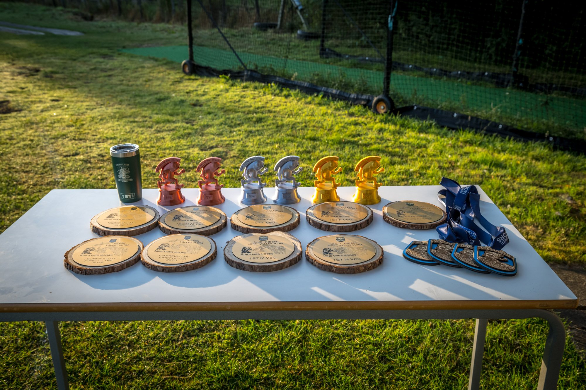 Table displaying several wood coasters with engraved text, multiple small race trophies in various colors, medals, a metal tumbler, and lanyards, set outdoors on a grassy area.