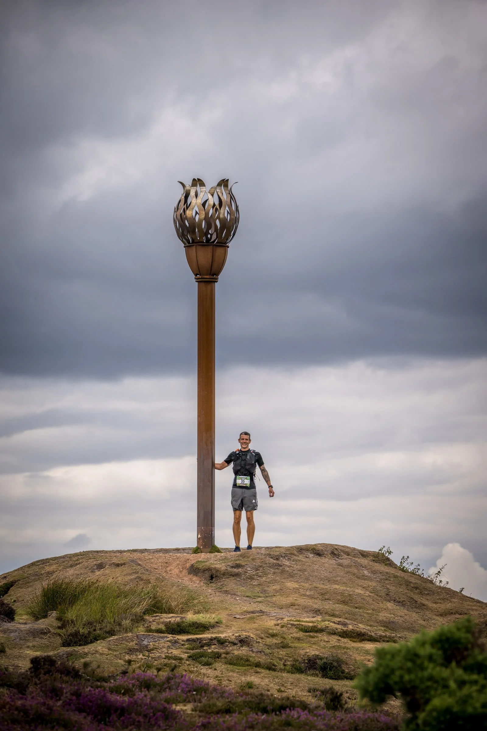 A person standing on a hill holding a tall, decorative torch with a cloudy sky in the background.
