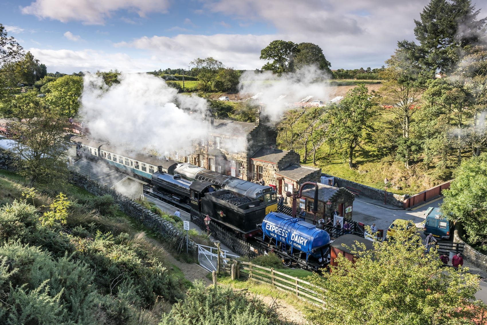 A vintage steam train with black engine and blue milk tanker car arriving at a station amidst trees and greenery, with smoke billowing from the engine.