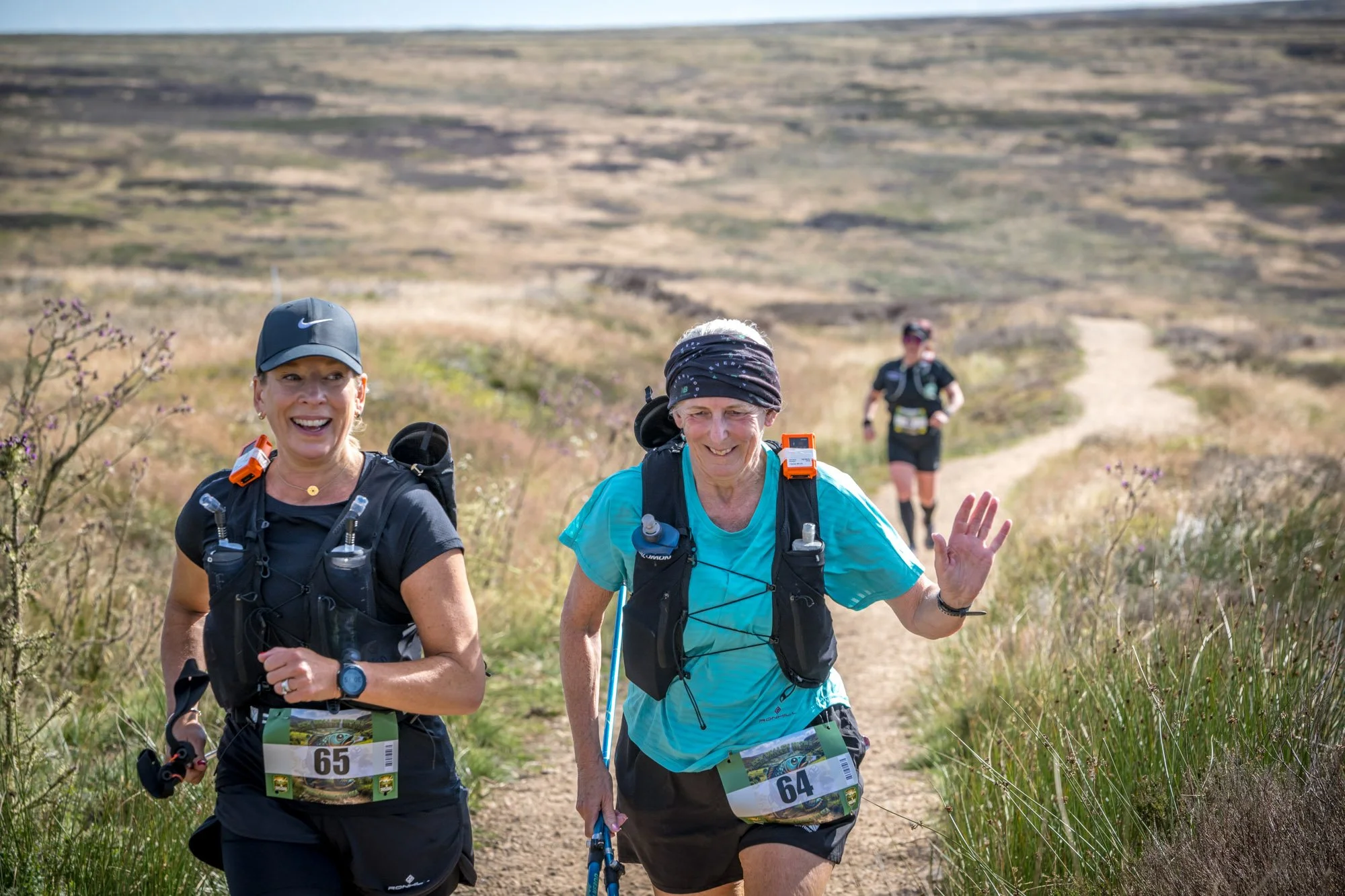 Two women running on a trail in a rural, grassy area, with a man in the background, all wearing athletic gear and race bibs, smiling and waving.
