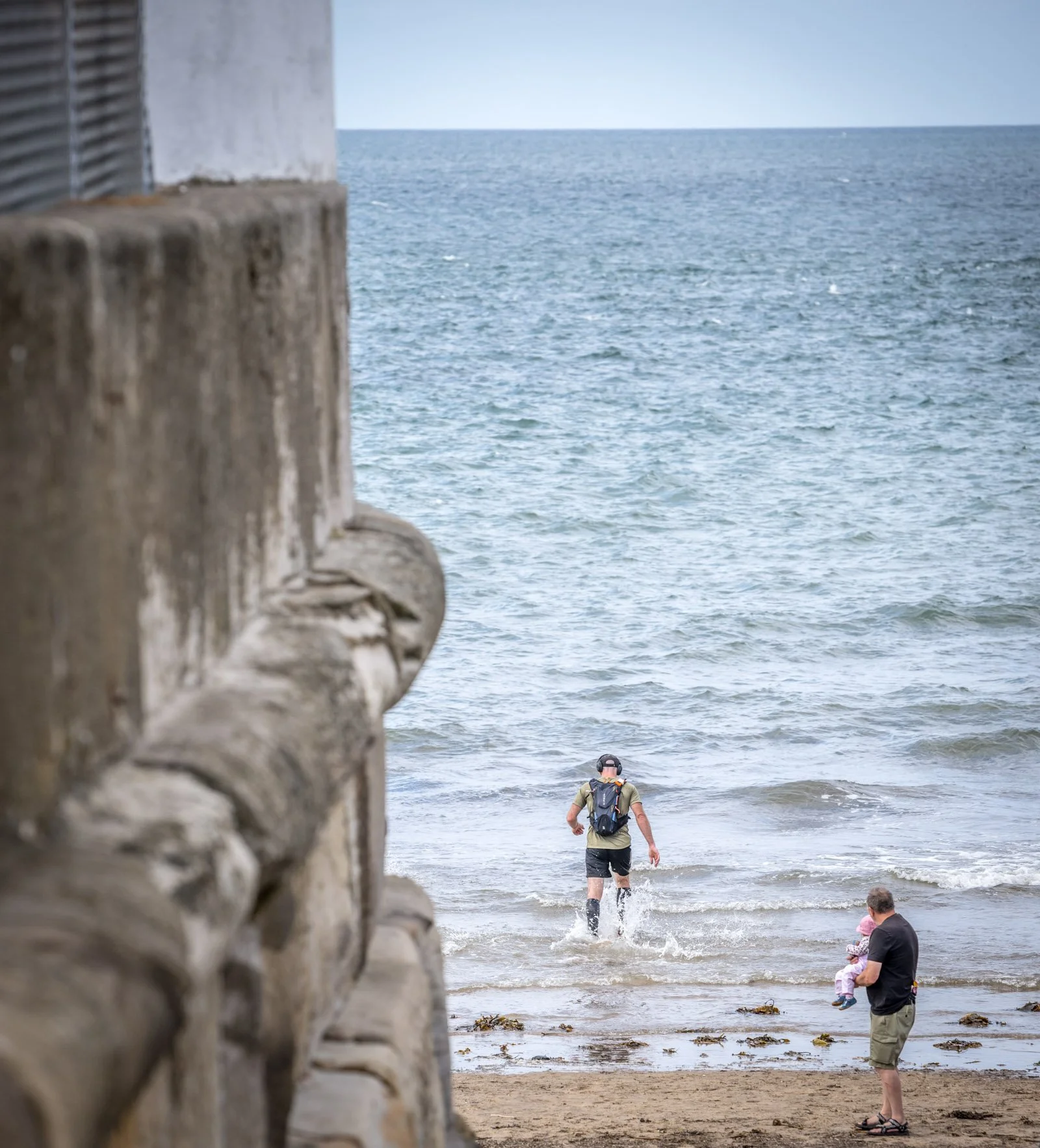 A person with a backpack walking out of the water onto the beach, with a child and an adult standing on the sand, seen near a rocky pier with the ocean in the background.