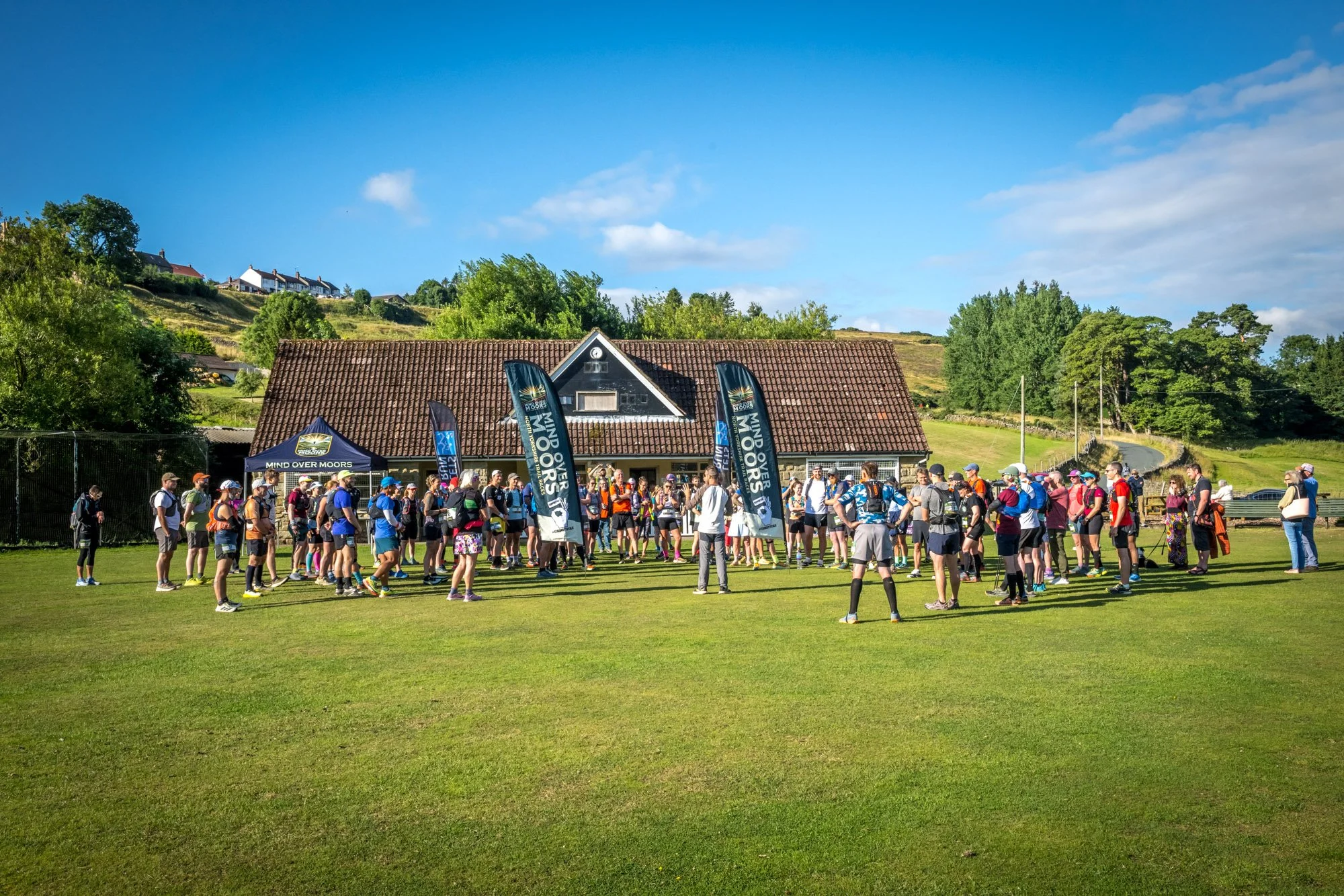 A group of runners gathered on a grass field, preparing for a race, with banners that say 'Mind Over Moors' and a building with a brown tiled roof in the background, under a blue sky with some clouds.