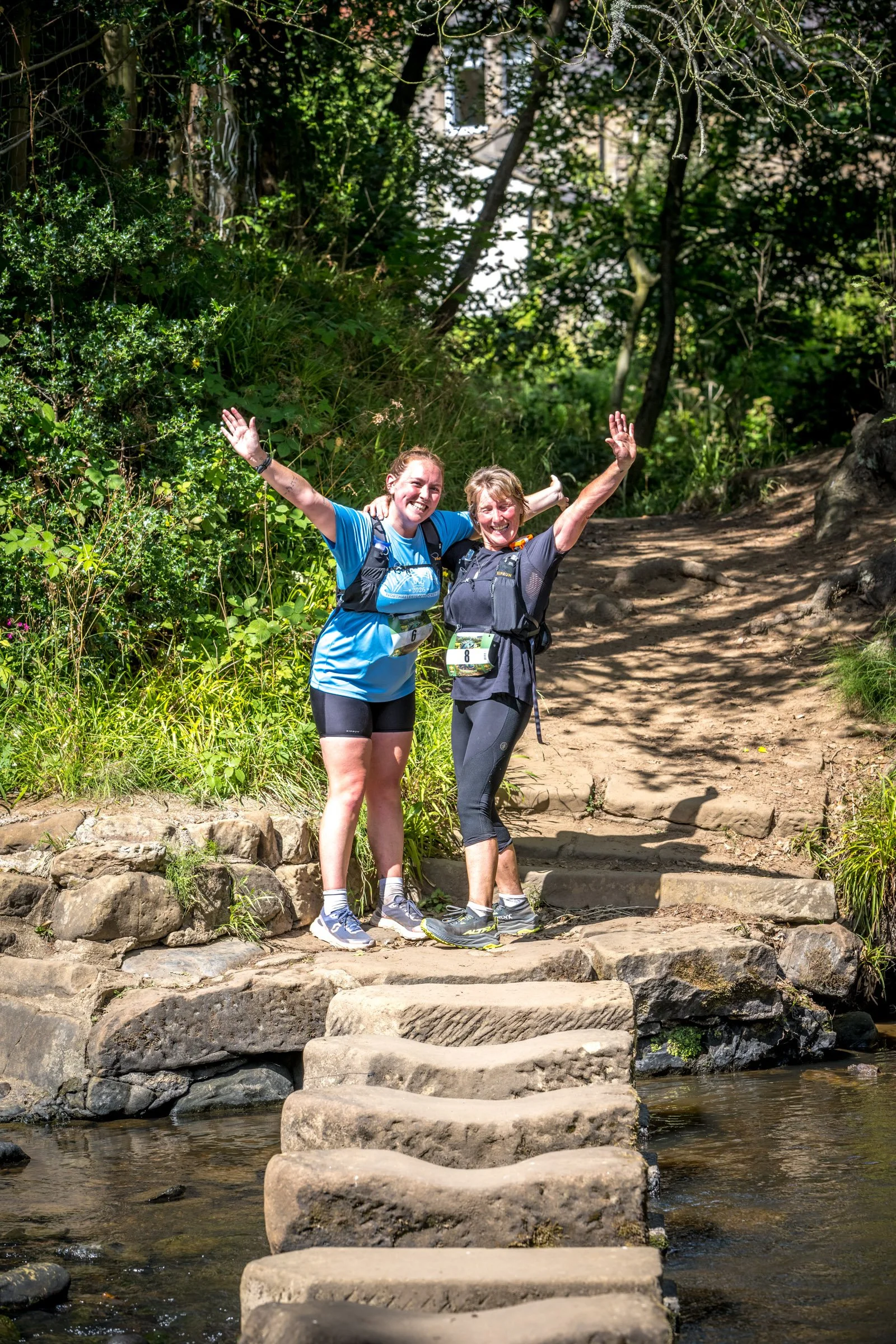 Two women with race bibs and hydration packs smiling and celebrating on a stone stairway crossing a small stream in a wooded area.