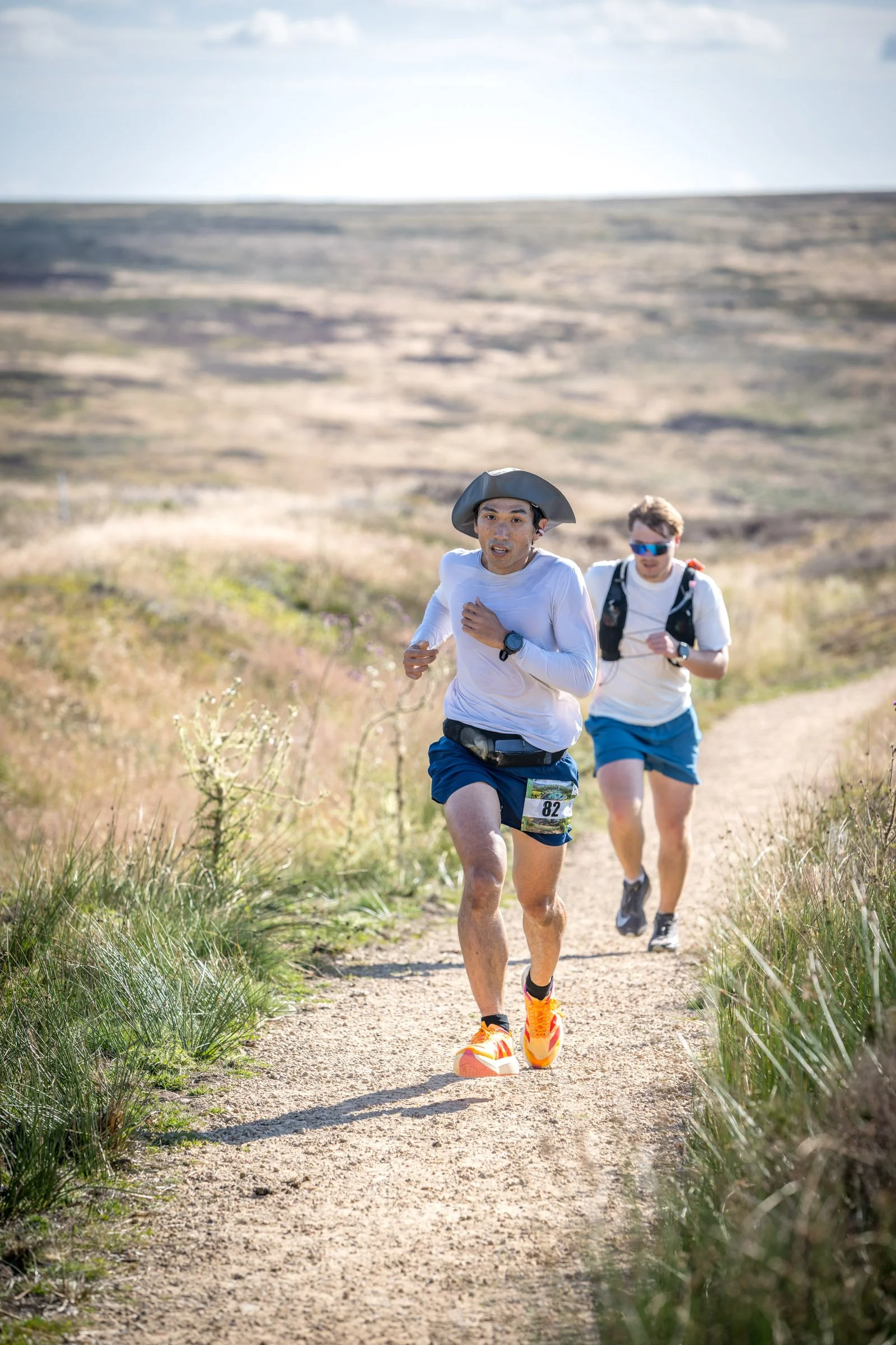 Two male trail runners running on a dirt path in a grassy field, with rolling hills in the background. One runner is wearing a hat, white shirt, and blue shorts, while the other is wearing sunglasses, a white shirt, and blue shorts.