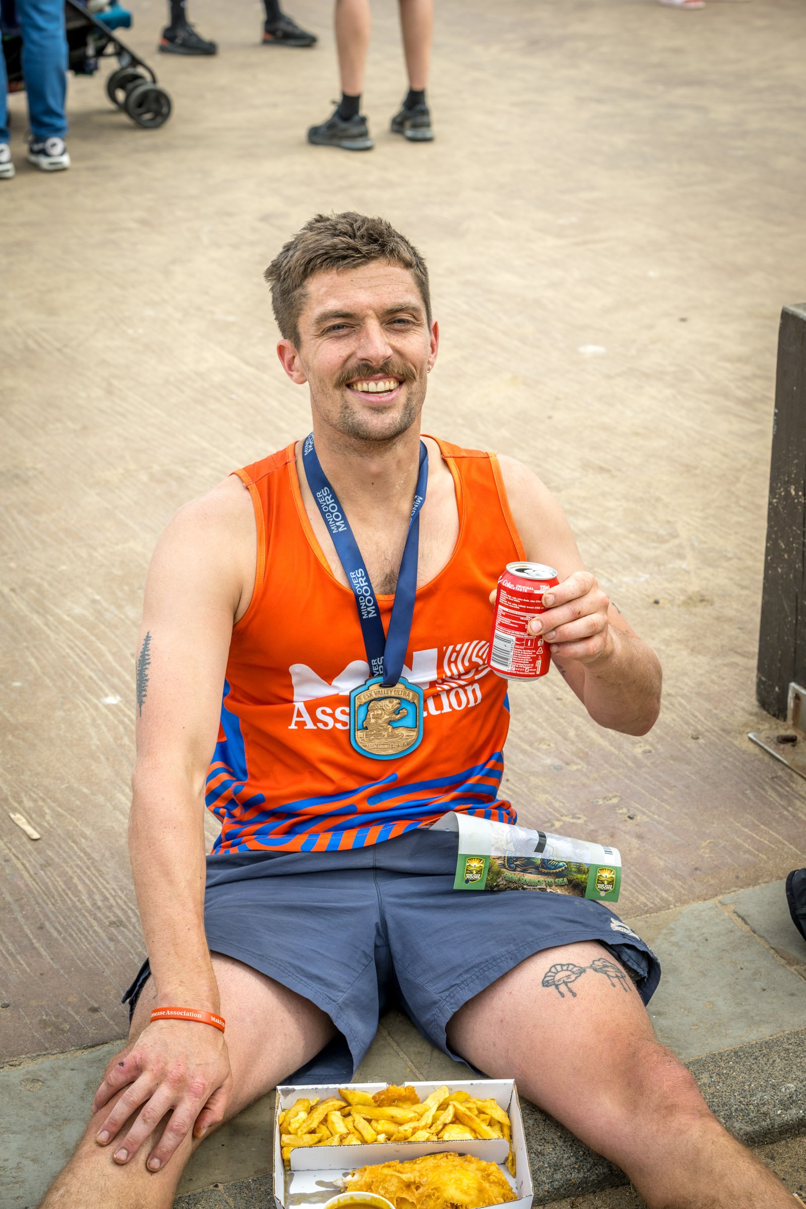 A smiling man wearing an orange tank top and gray shorts, sitting on the ground with a medal around his neck and holding a can of soda, is enjoying a meal of fries and battered fish in a box. There are other people and a stroller in the background of