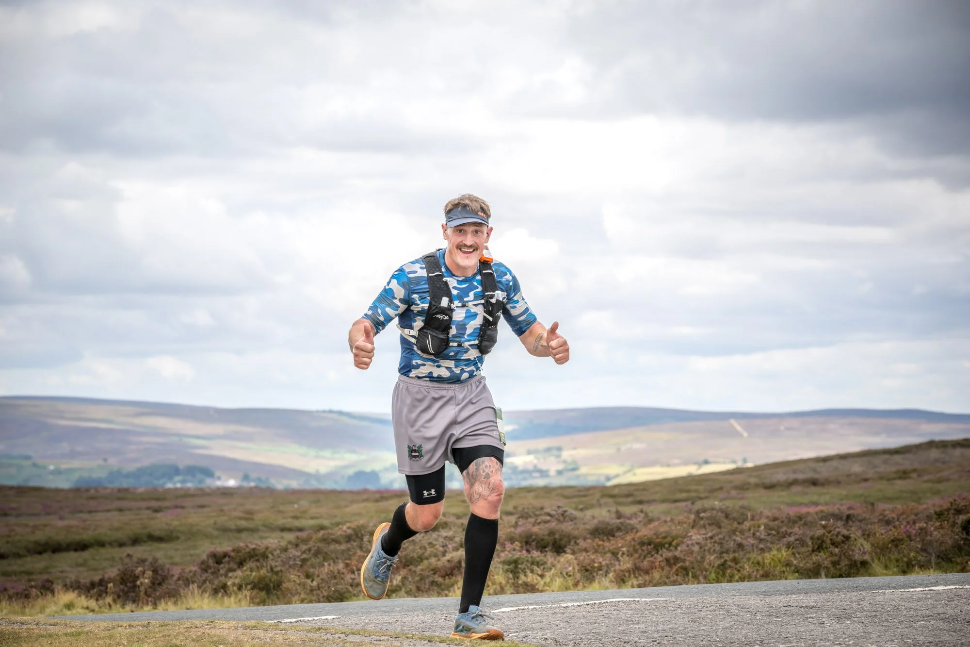 A man dressed in athletic gear running outdoors on a cloudy day, with a landscape of open fields and rolling hills in the background.