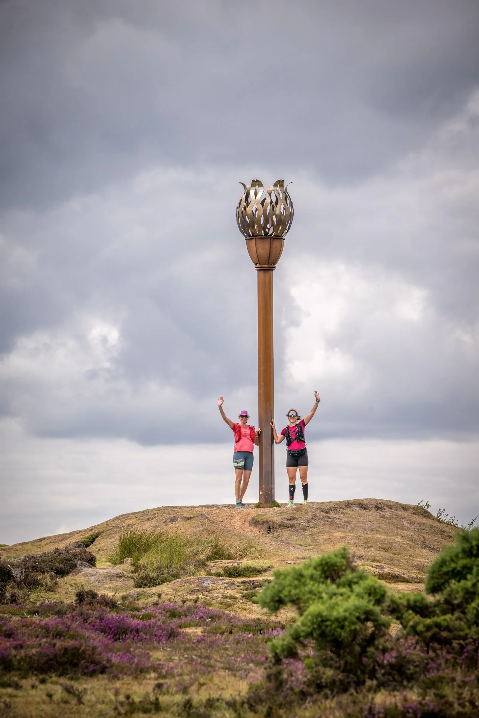 Two women in athletic clothing and backpacks standing on a hilltop next to a tall decorative metal torch with a cloudy sky behind them.