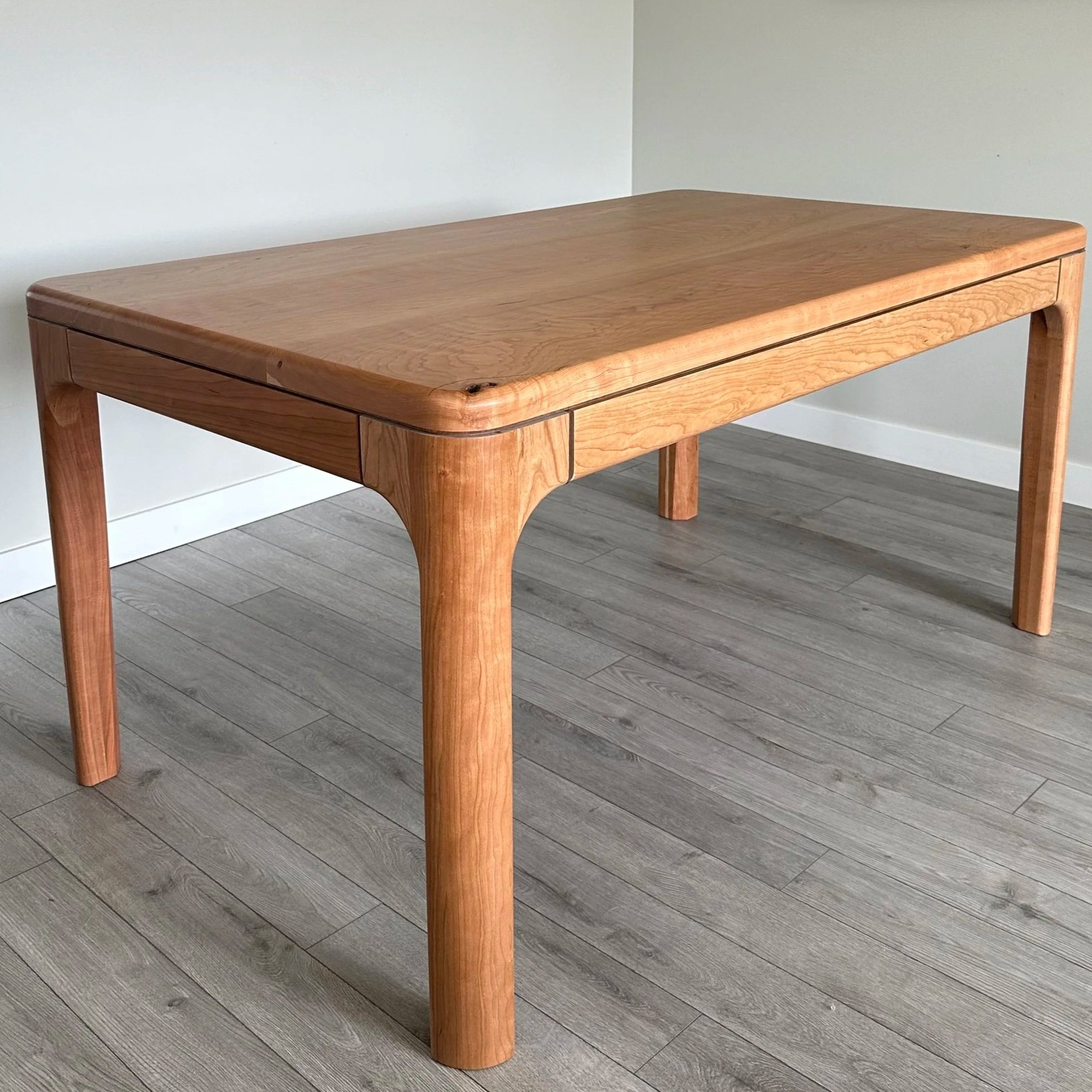 A cherry wood table with a rectangular top and curved, sturdy legs, set on a gray wood floor against a white wall.