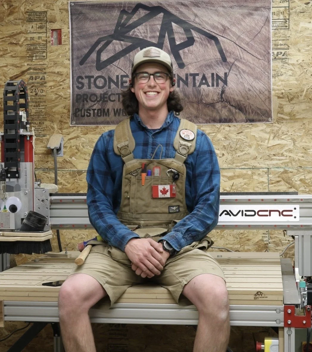 A cover photo of Harley Griffin Owner of the Calgary based Woodworking shop Stone Mountain Projects  posing with a big smile with his work vest on sitting on his cnc machine with his logo flag pinned up behind him