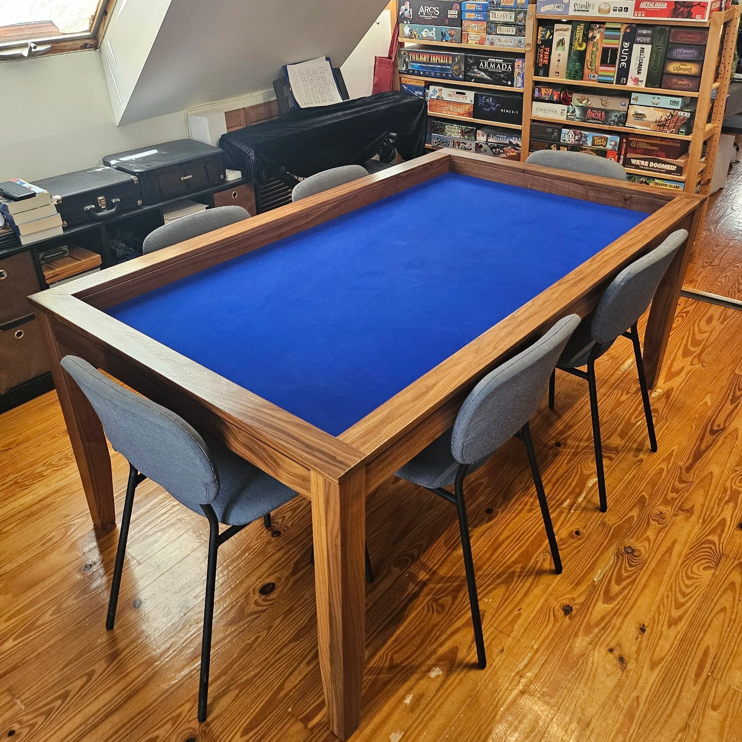 Corner view of a sleek walnut game table with grey chairs tucked in and a blue neoprene playing surface