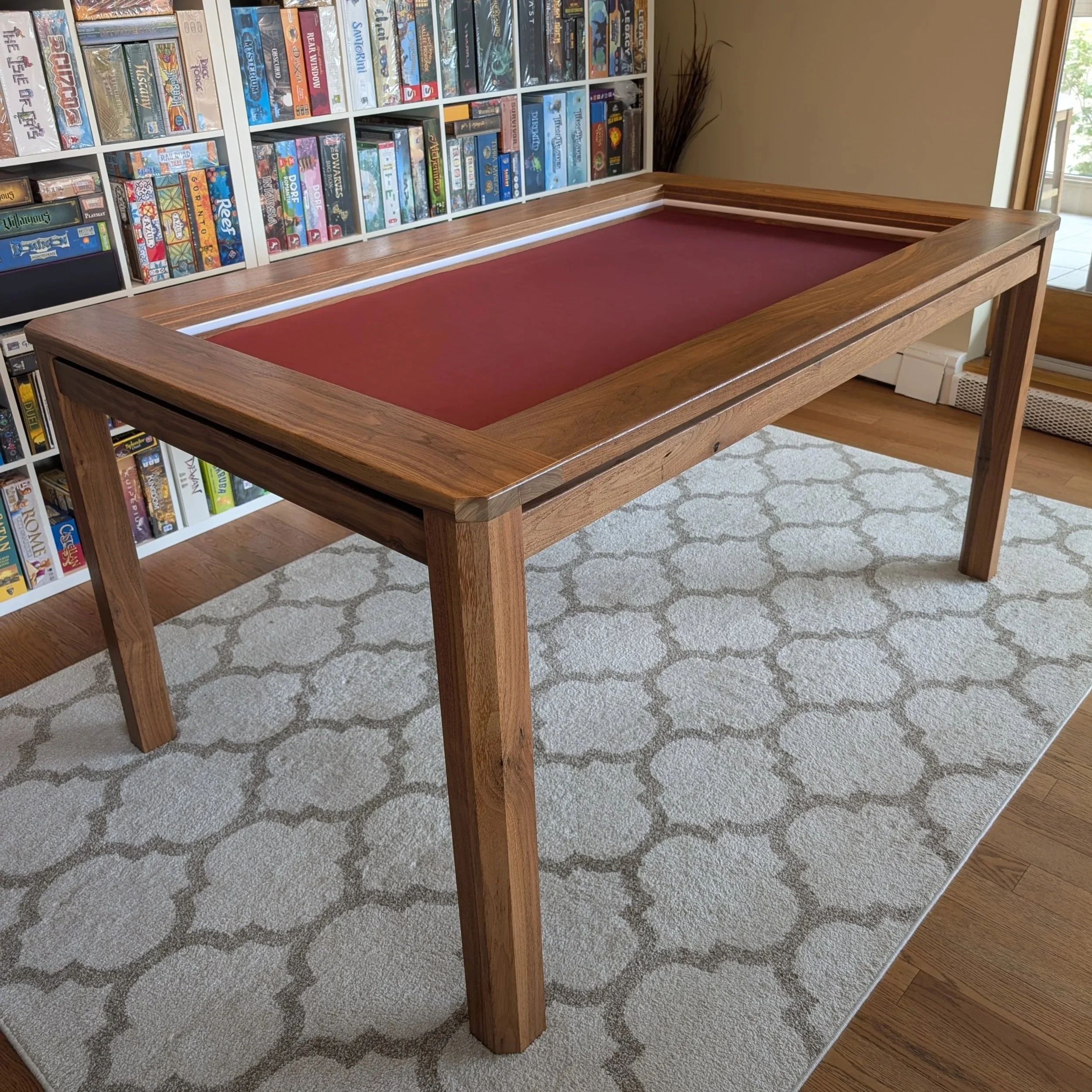 Walnut board game table with a recessed red neoprene playing surface, shown in a room with a board game collection on shelves behind it. Handcrafted in Calgary, Alberta by Stone Mountain Projects.