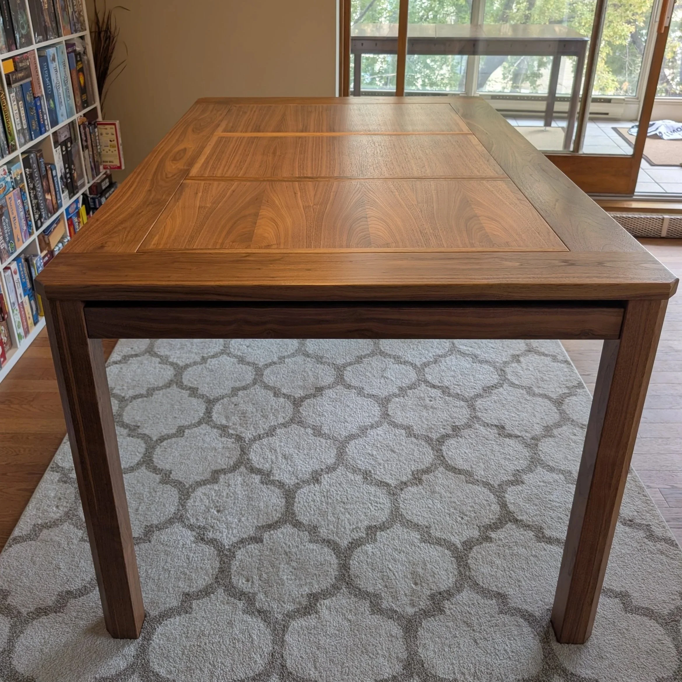 Front view of a walnut board game table showing its tapered legs and paneled tabletop, with a board game shelf and window in the background.