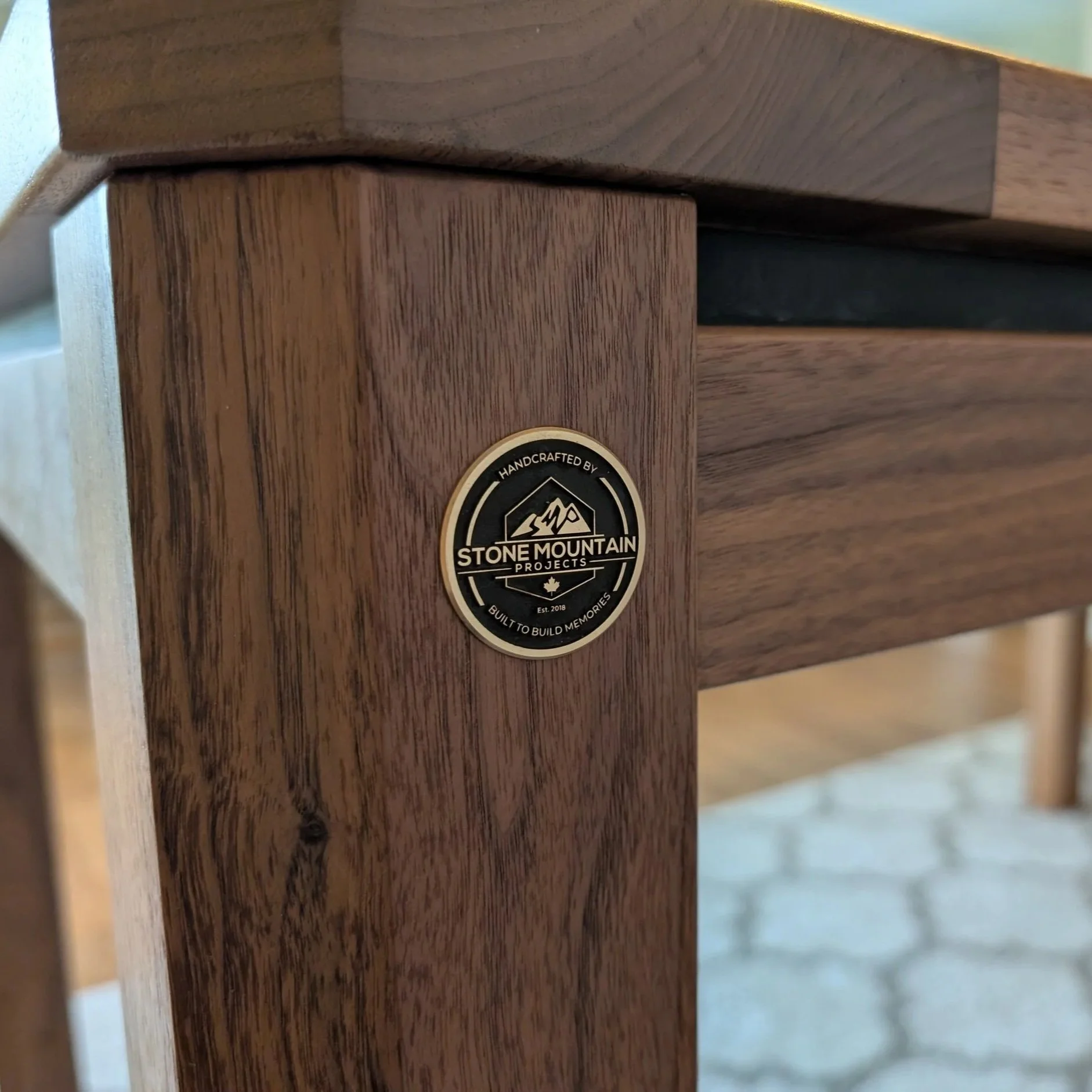 Brass "Handcrafted by Stone Mountain Projects" badge inlaid into a walnut table leg, showing the joint between two-tone walnut grain. Built in Calgary, Alberta by Stone Mountain Projects.