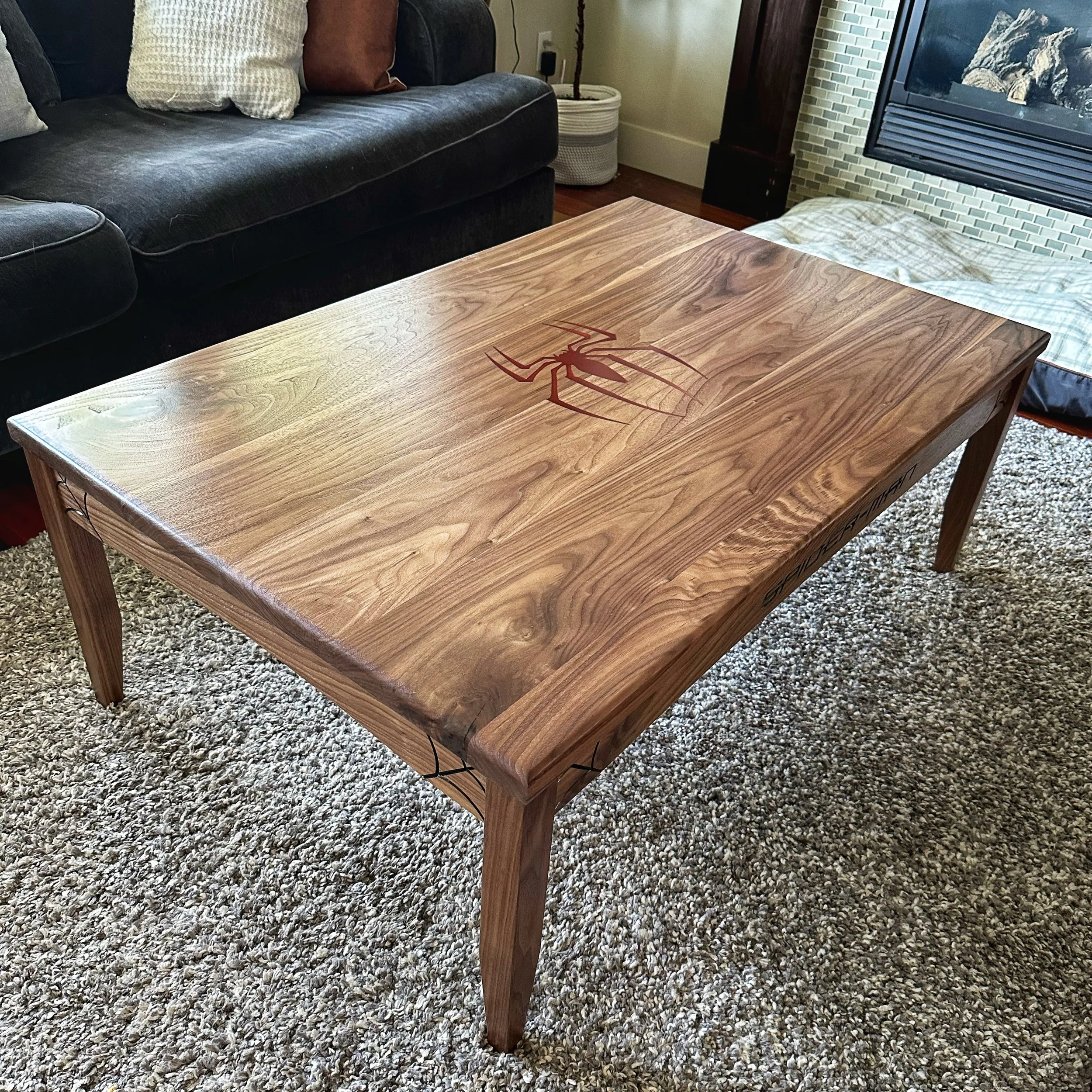 A solid walnut coffee table with a Spiderman logo engraved into the middle of it and cobweb accents in the corner of the aprons