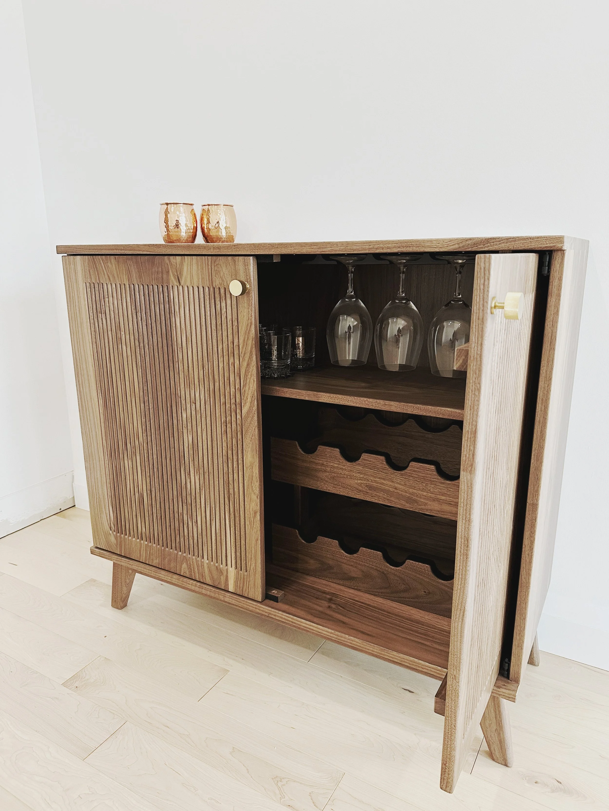 A walnut dry bar with fluted front doors one door is open revealing the inside wine rack and glass holder
