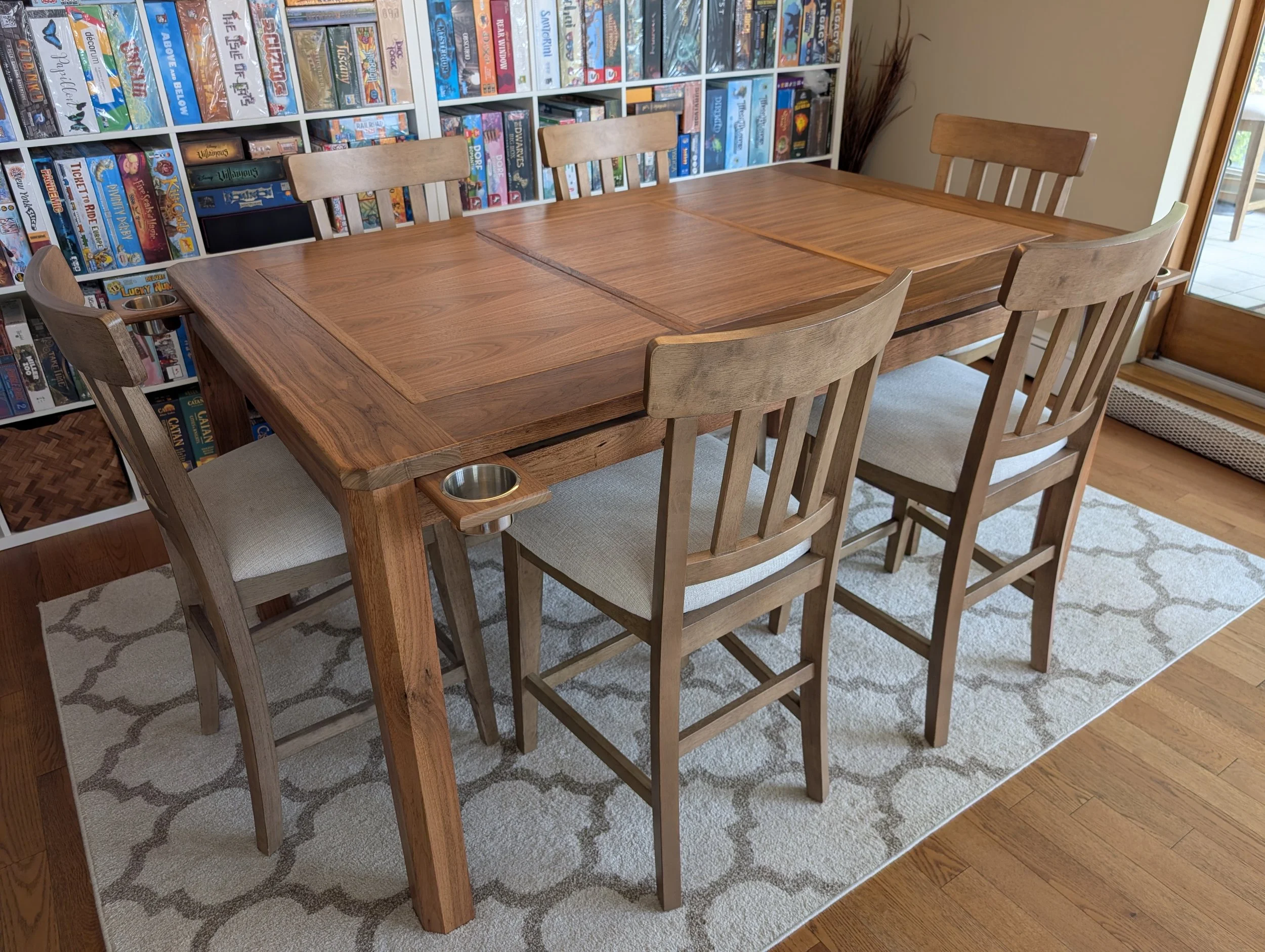 view from the corner of a finished walnut board game table with six chairs, magnetic cupholders at the corners, and a board game collection on shelves behind it. Handcrafted in Calgary, Alberta by Stone Mountain Projects.