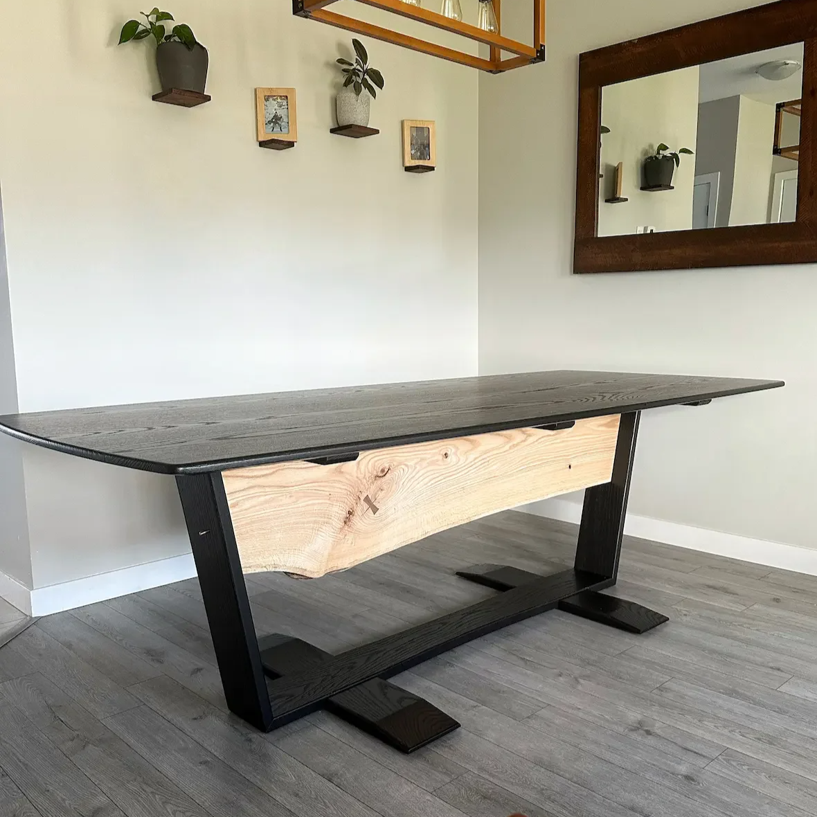 A modern dining table with a  black frame  and top in a room with light-colored walls, a large mirror, and small wall-mounted planters and framed pictures.