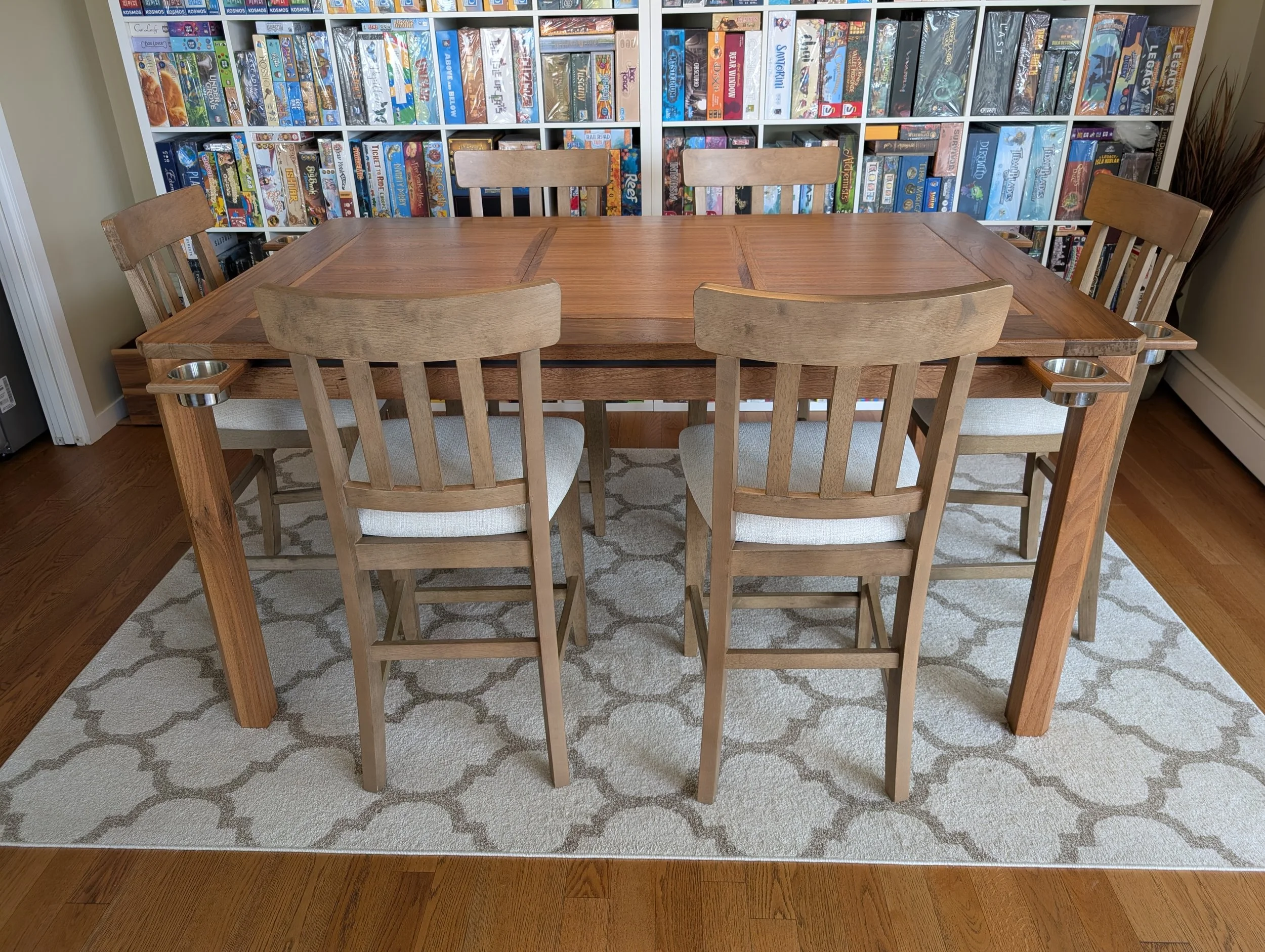 Side view of a finished walnut board game table with six chairs, magnetic cupholders at the corners, and a board game collection on shelves behind it. Handcrafted in Calgary, Alberta by Stone Mountain Projects.