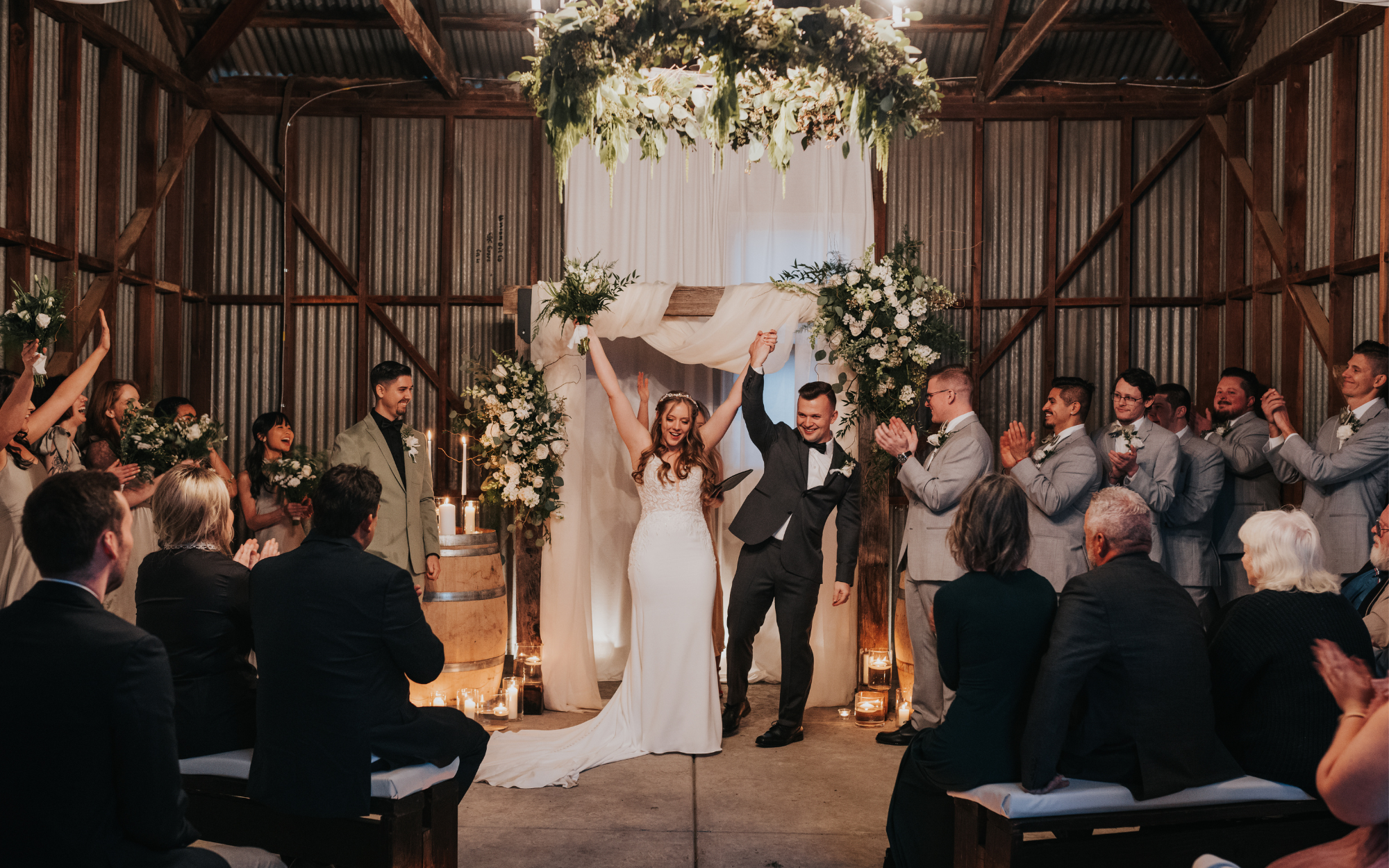 Bride and groom celebrate their wedding with friends and family in a rustic barn, surrounded by floral arrangements and candles.