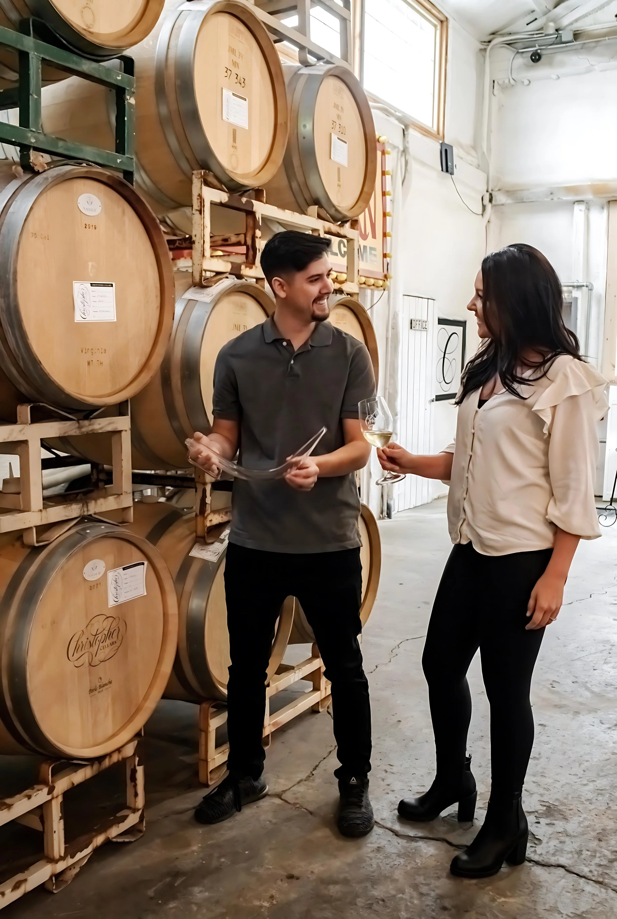 Christopher Mora smiling as he thieves Chardonnay from the barrel and pours Candice Mora a taste, sharing a quiet moment of evaluation and trust in the cellar.