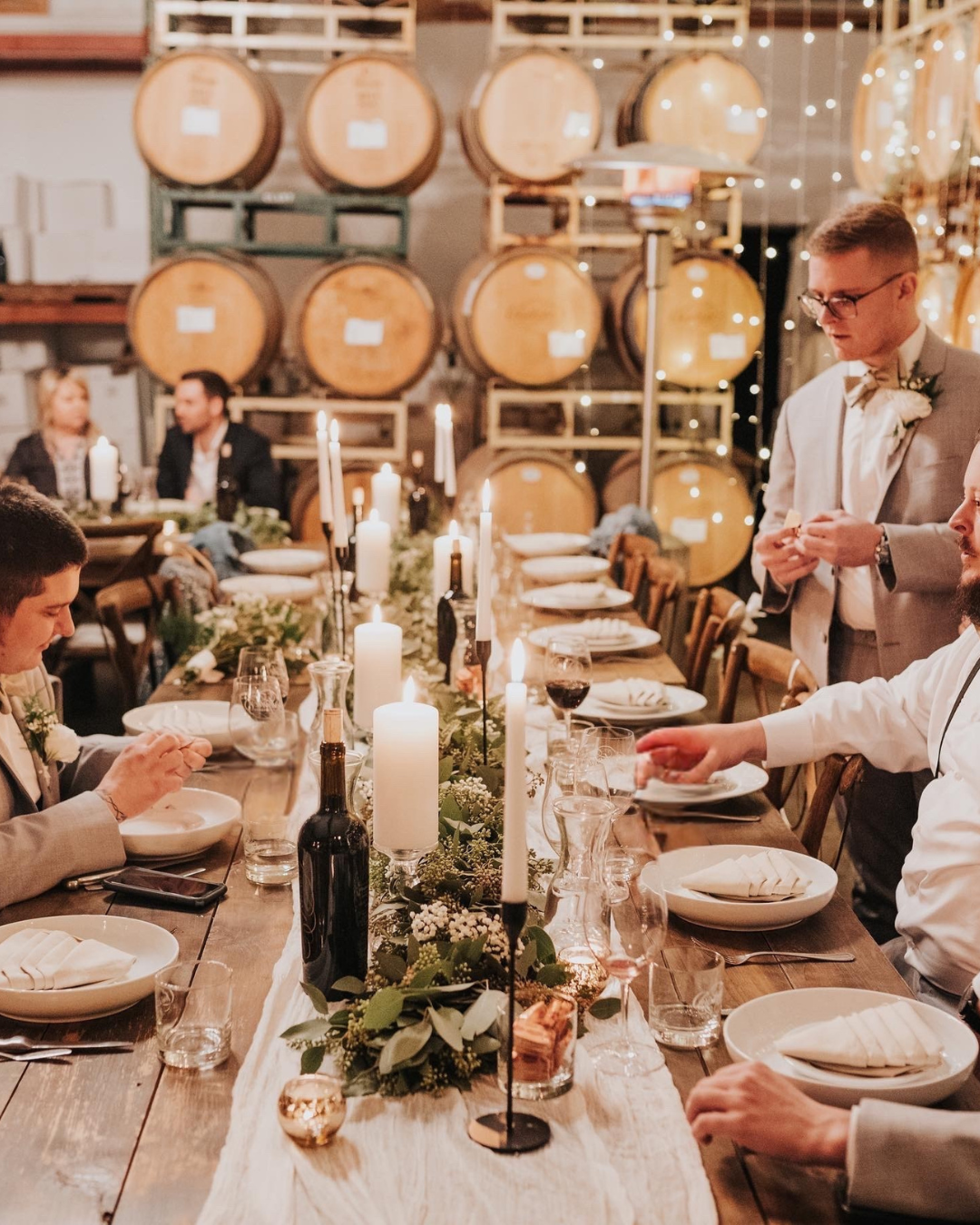 A formal dinner party set in a rustic venue with wine barrels, candles, and floral decorations. Guests are dressed in semi-formal attire, with some standing and some seated at a long wooden table.