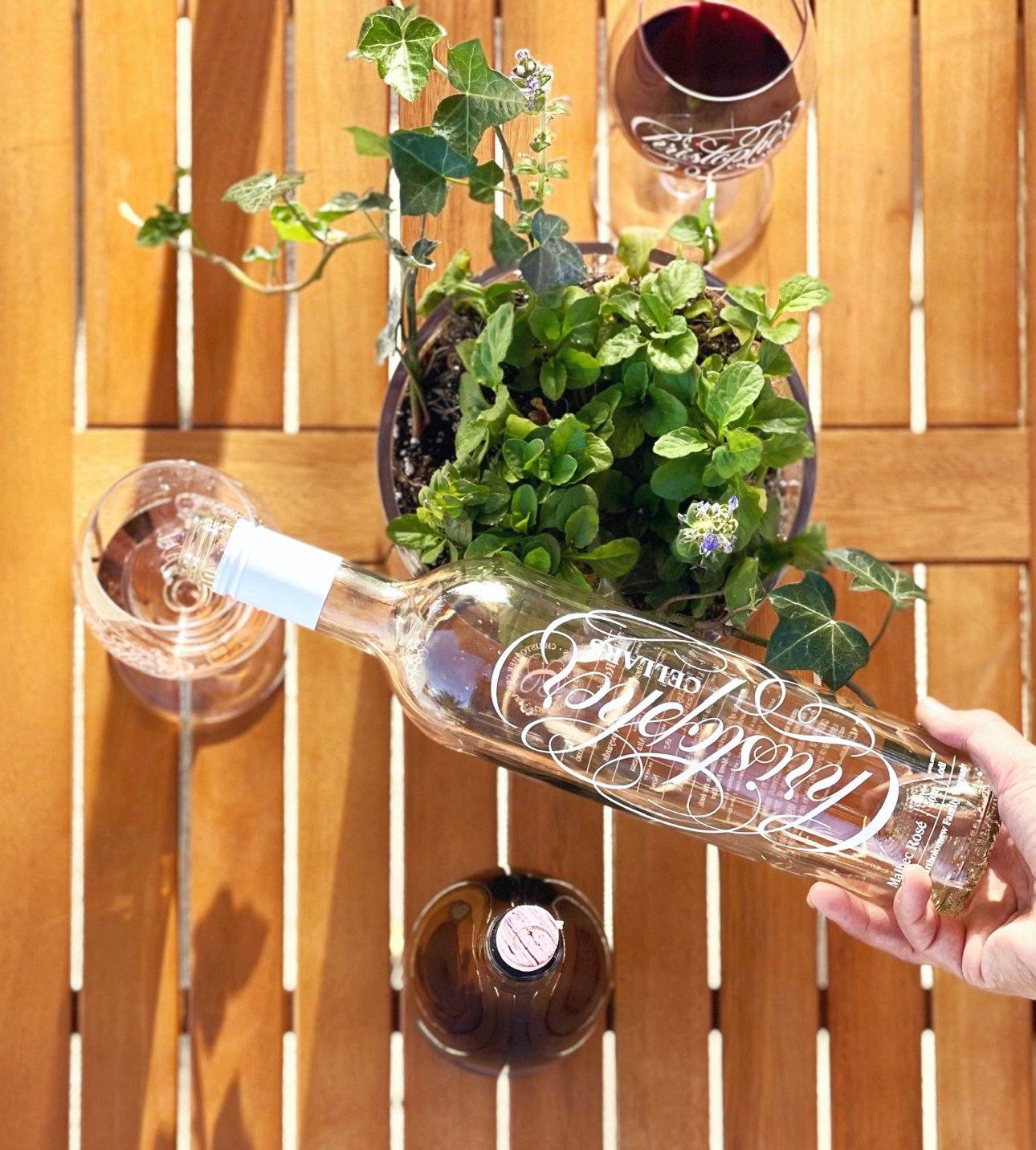 A top-down view of a wooden table with a potted plant, two glasses of wine, an empty small bottle, and a hand holding a large bottle of rosé wine.