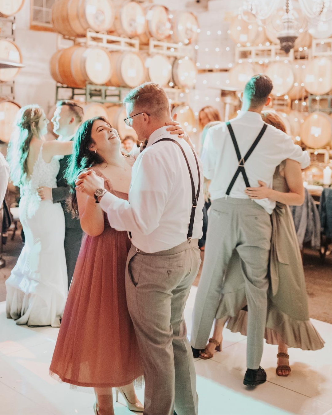A couple dancing at their wedding reception at a winery, with oak barrels and string lights creating a romantic atmosphere.
